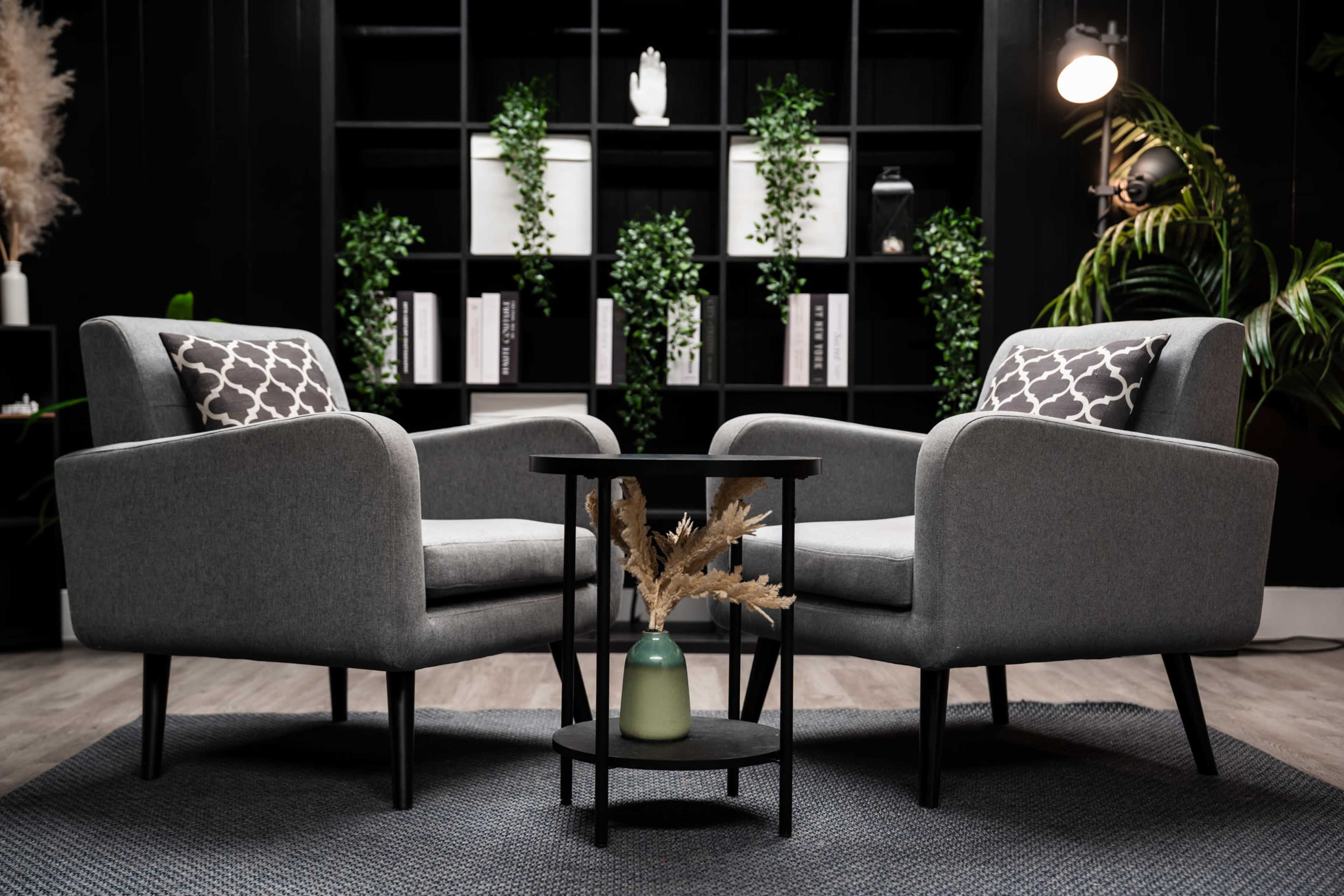 Two gray armchairs are positioned across from each other around a small, round black table with a decorative vase and dried plants, set against a black bookshelf filled with green plants and white decor.