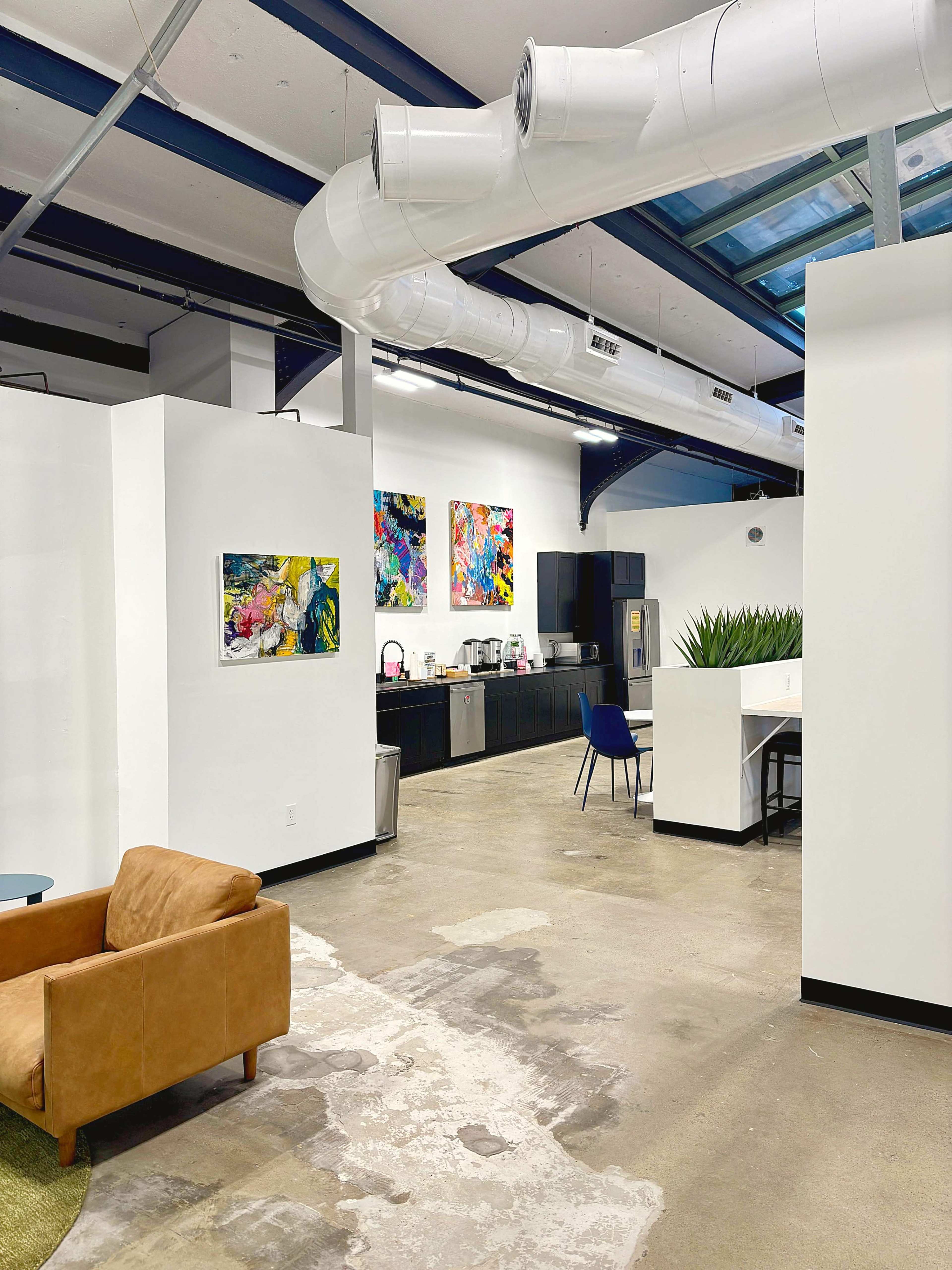 The image shows a modern office space featuring a seating area with a brown couch, a kitchenette with dark cabinets, and colorful artwork on the walls.