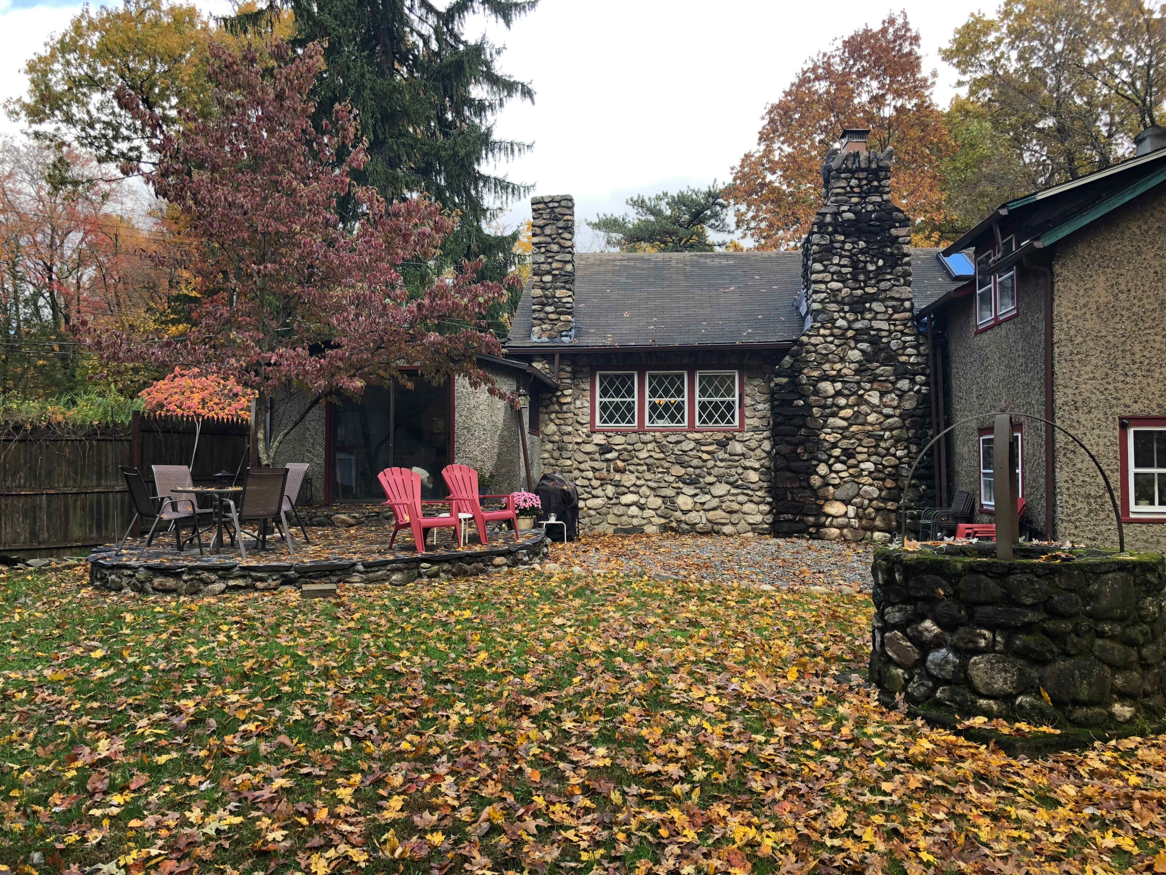 A stone cottage surrounded by trees and a yard covered in fallen leaves, featuring pink chairs and a circular stone fire pit.