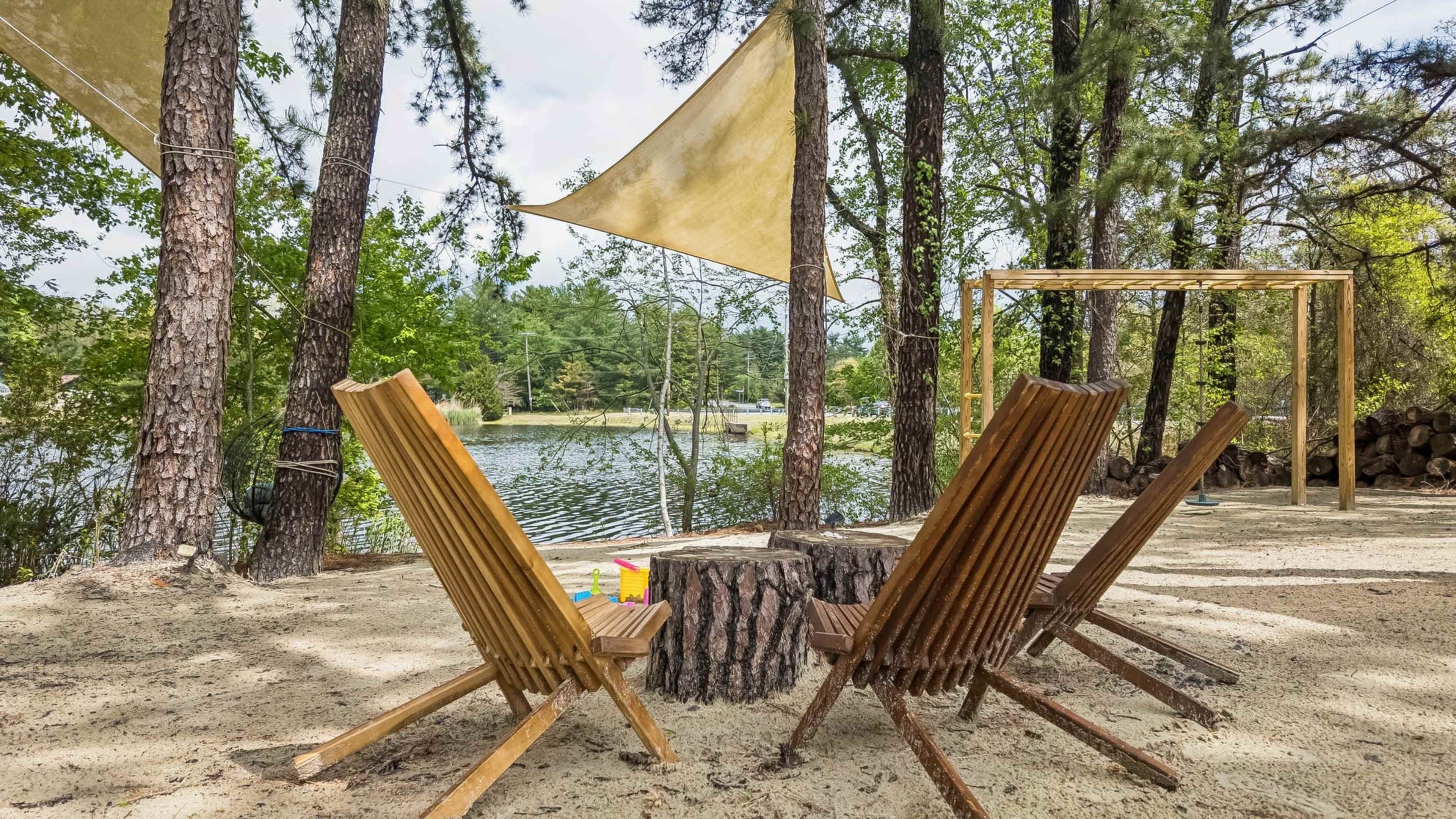 The scene features four wooden chairs arranged around a tree stump in a sandy area near a tranquil lake, shaded by trees and awnings.