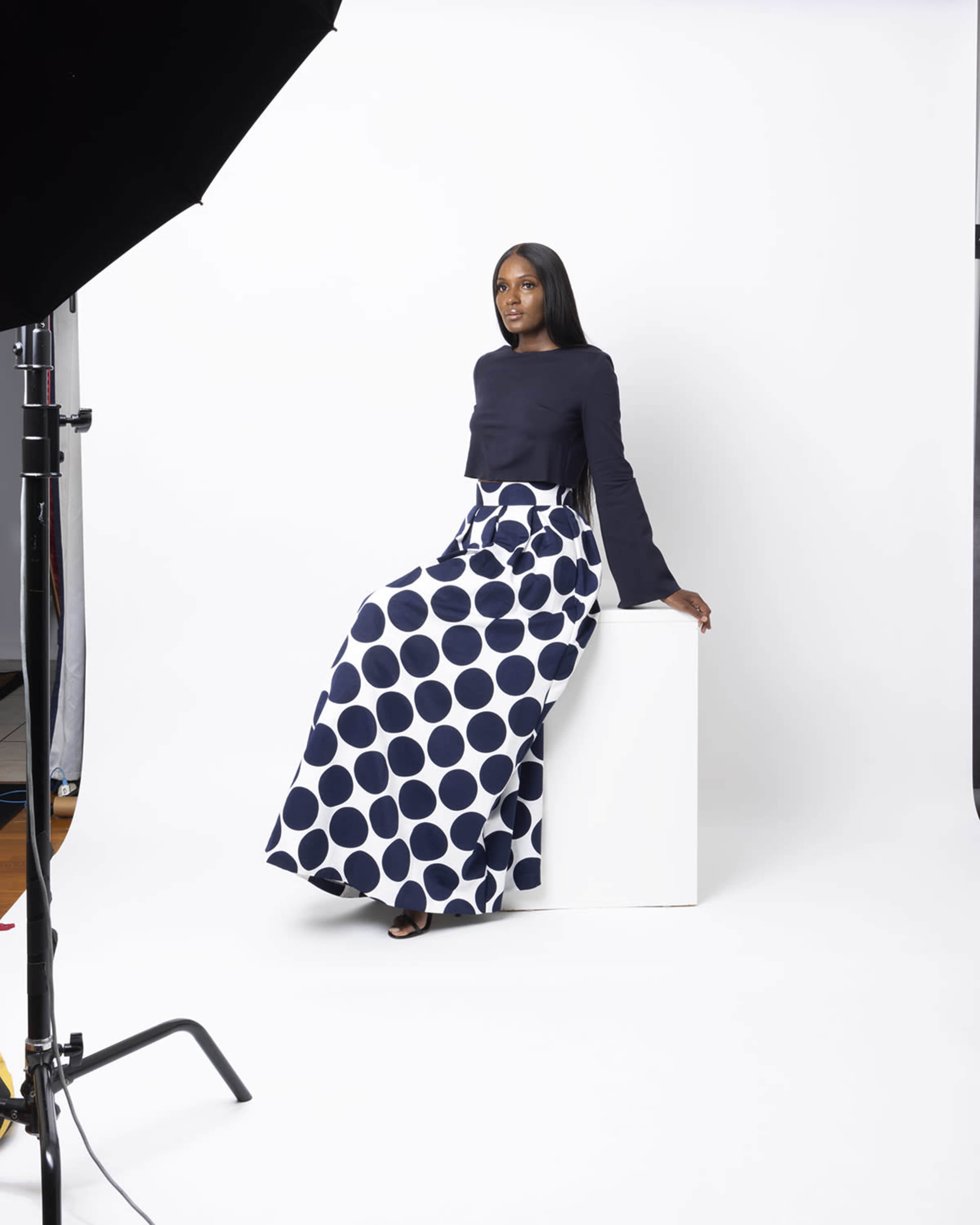 A woman sits on a white cubic pedestal, wearing a long, polka-dotted skirt and a solid-colored top, against a backdrop of a white studio.
