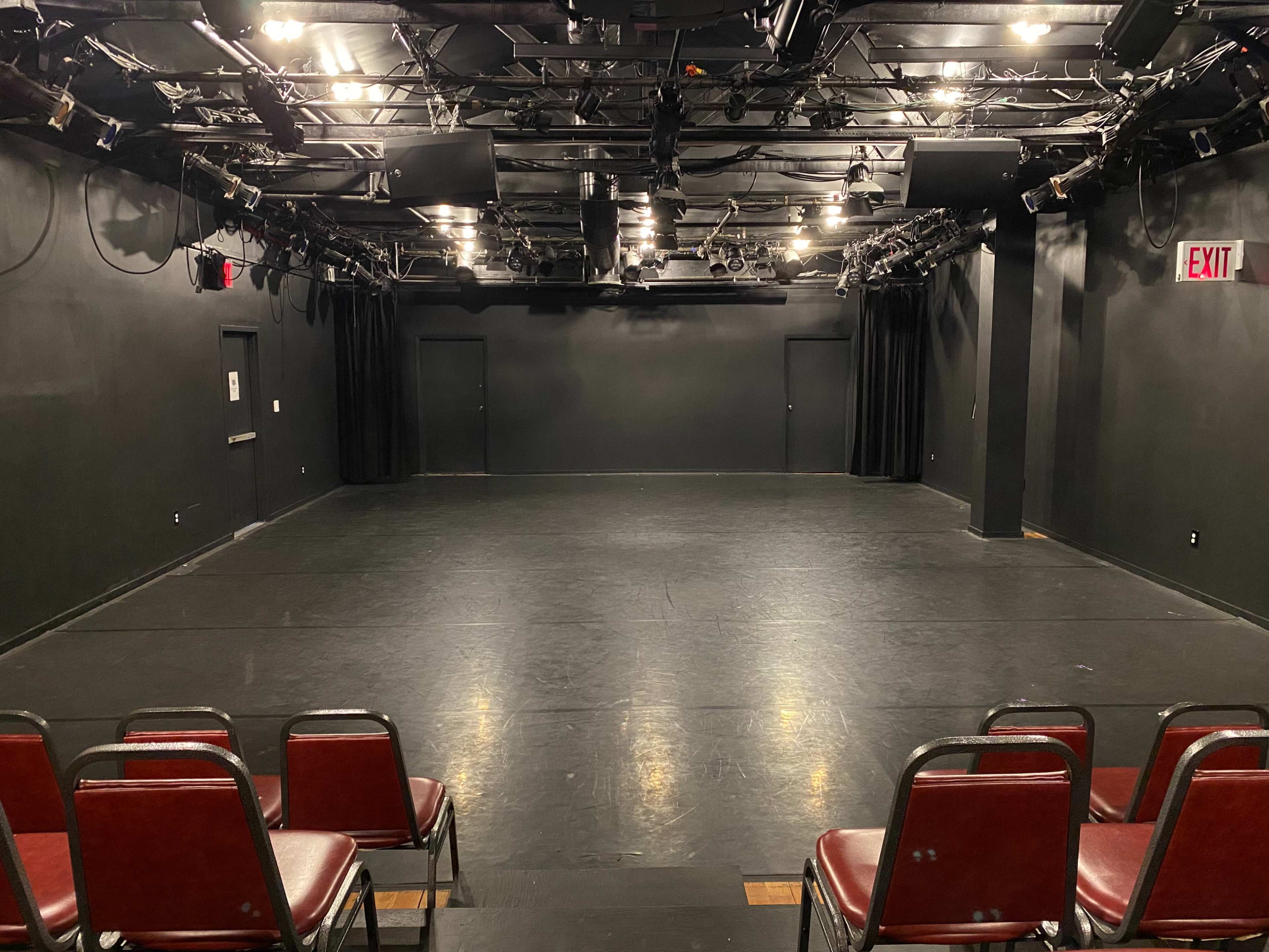 A dark theater stage with empty red chairs arranged in front, illuminated by overhead lights.