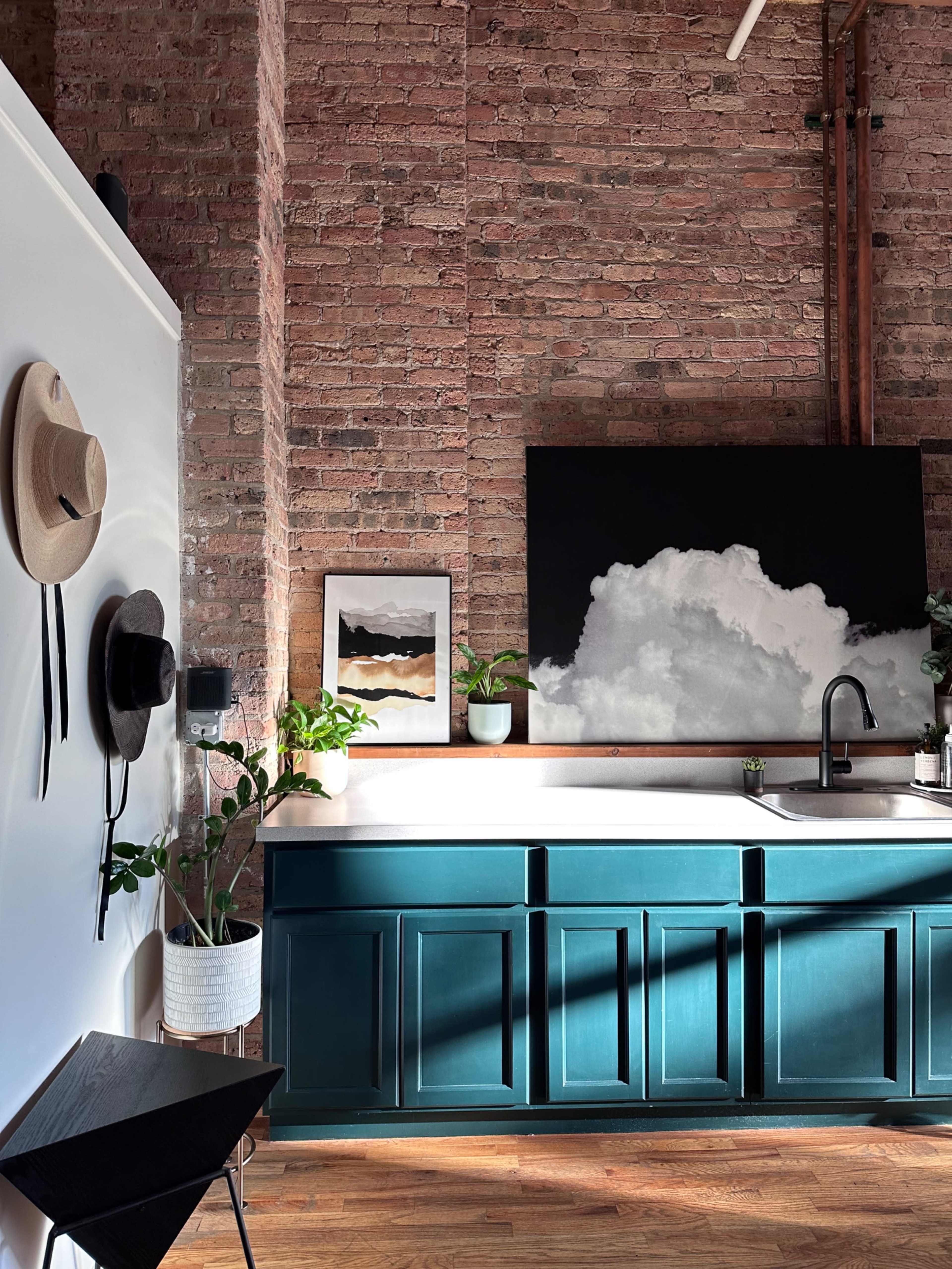 The image shows a modern kitchen with a deep teal cabinet, a white sink, exposed brick walls, and wall-mounted hats and plants for decoration.