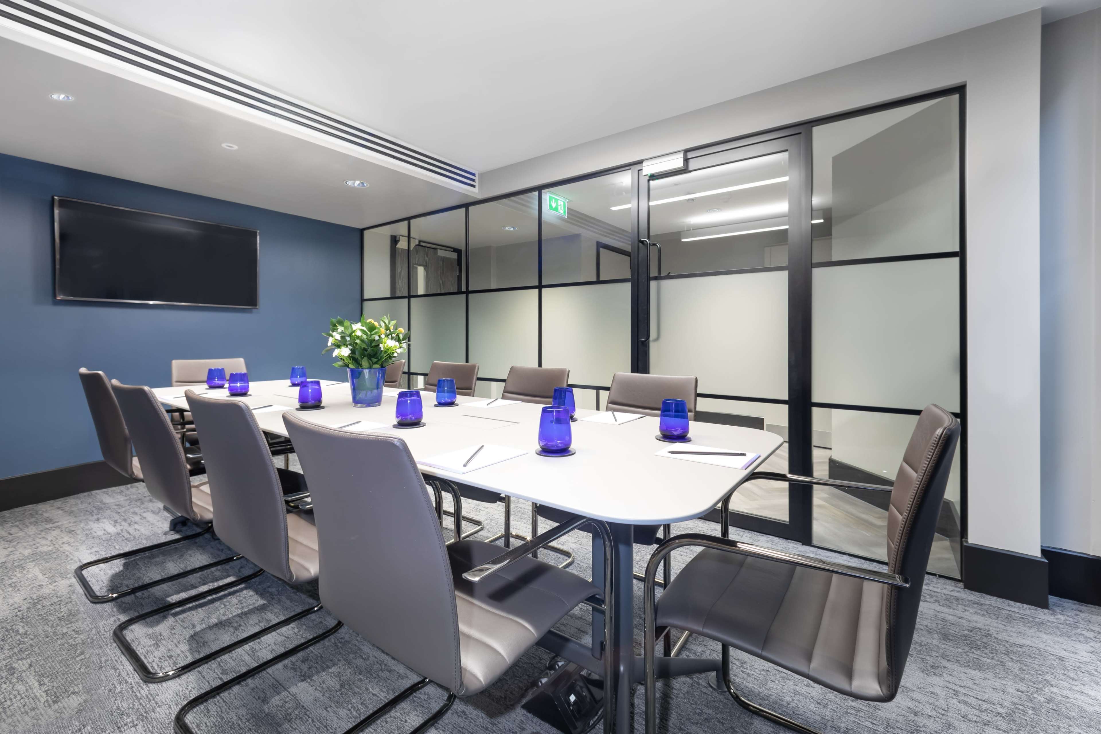 A modern conference room features a large table surrounded by eight ergonomic chairs, with a television mounted on one wall and glass partitions separating it from the hallway.