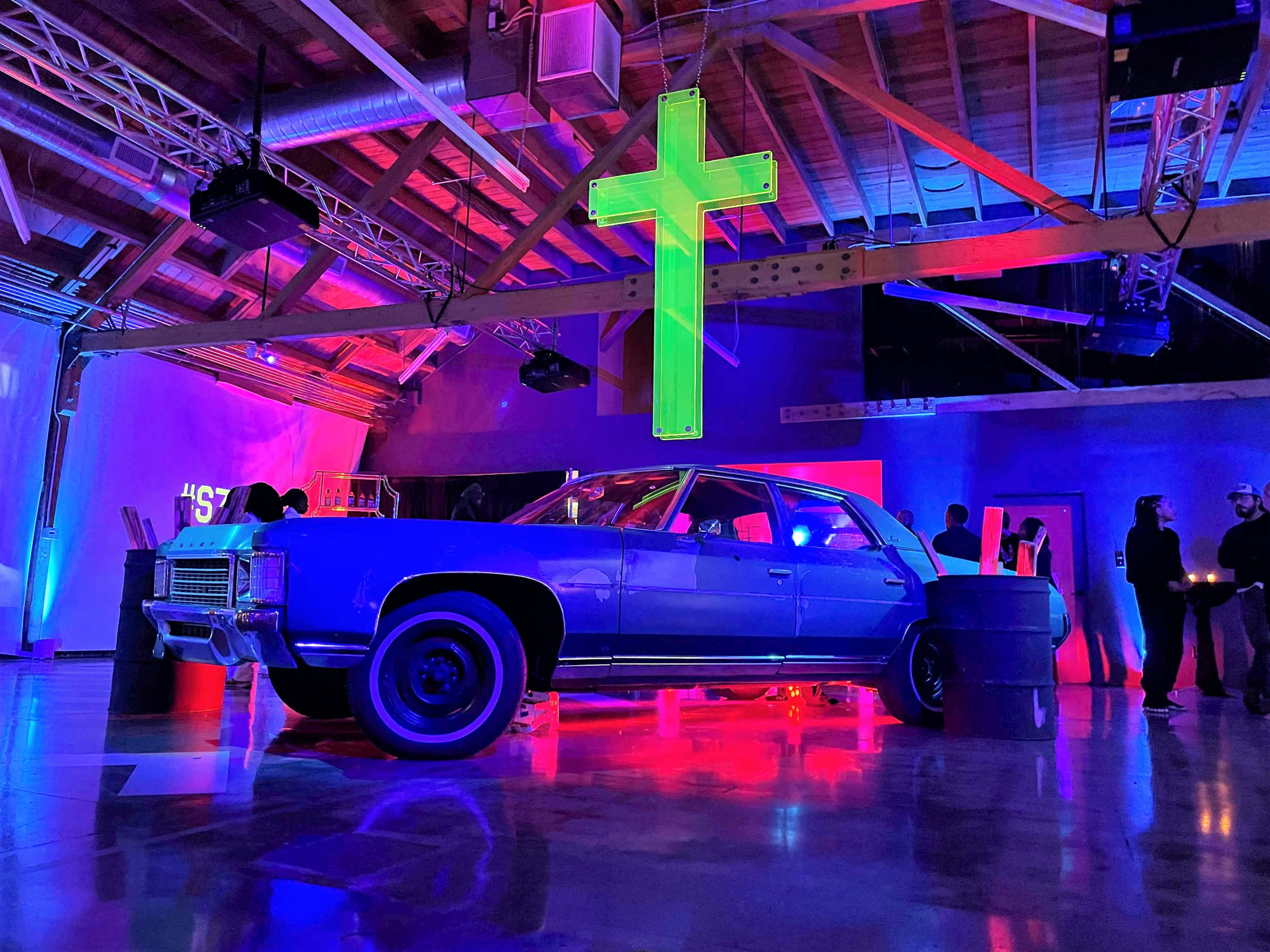 A blue classic car is positioned on a stage under colorful lighting with a large green cross hanging above it in a warehouse-like space.