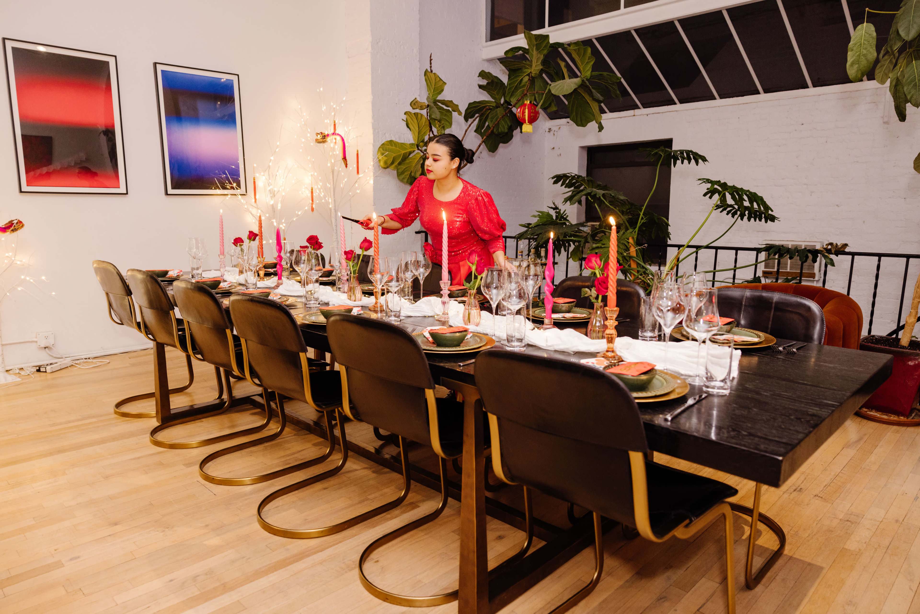 A woman in a red outfit sets a long dining table adorned with candles and floral centerpieces in a modern, well-lit room.