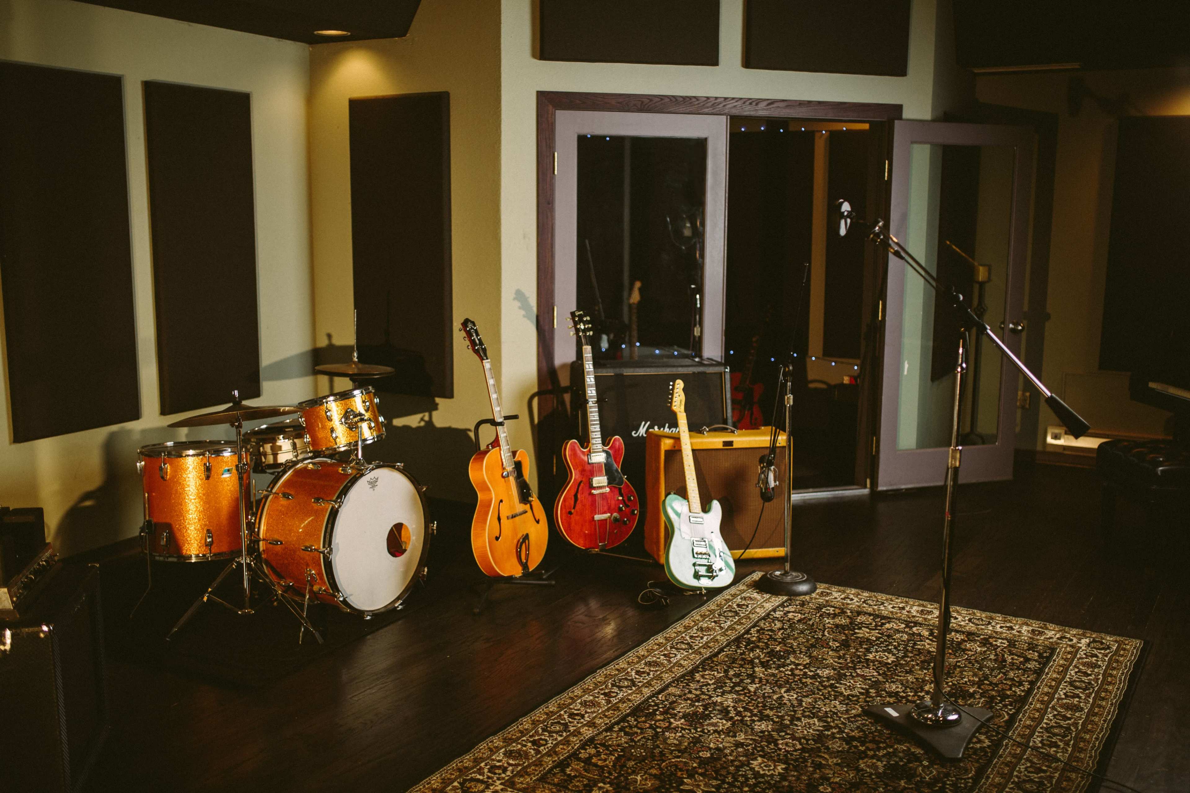 A music studio features a drum set, several guitars, an amplifier, and a microphone on a patterned rug.