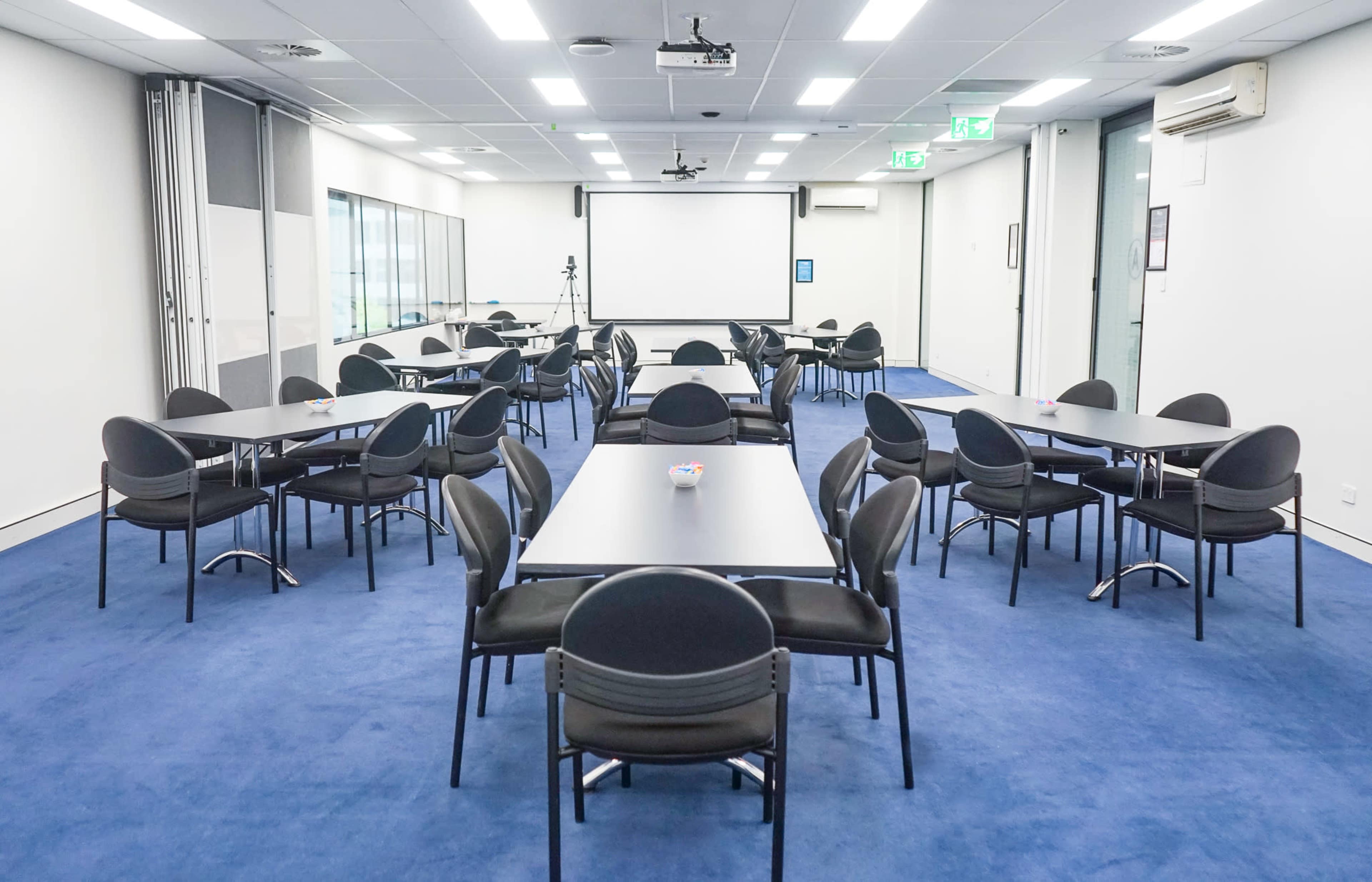 The image shows a professional meeting room with multiple tables and chairs arranged in a clean, organized layout on a blue carpet.