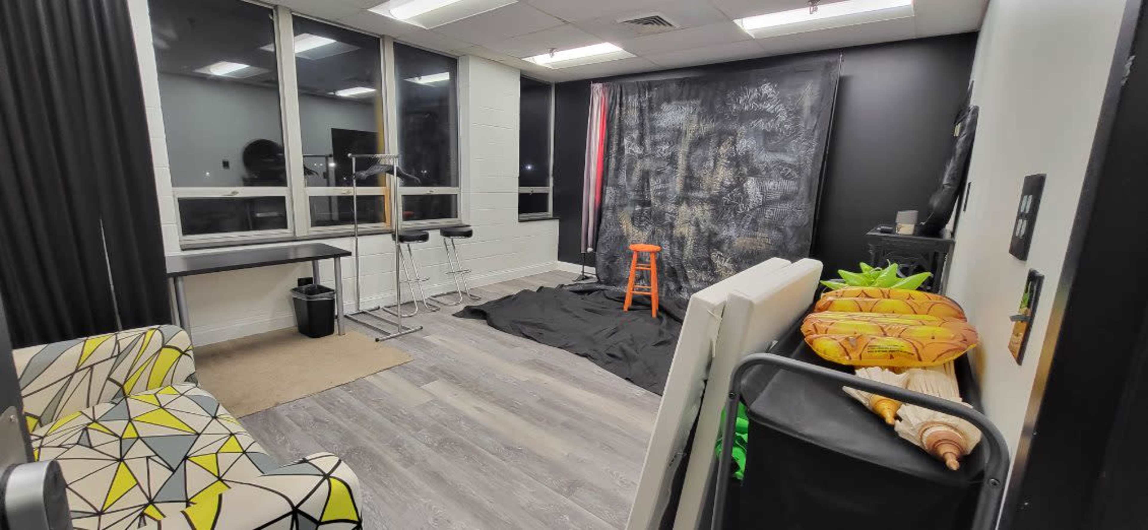 The photo shows a small studio room with a black backdrop, an orange stool, a patterned sofa, and equipment like a table and a cart in the space.