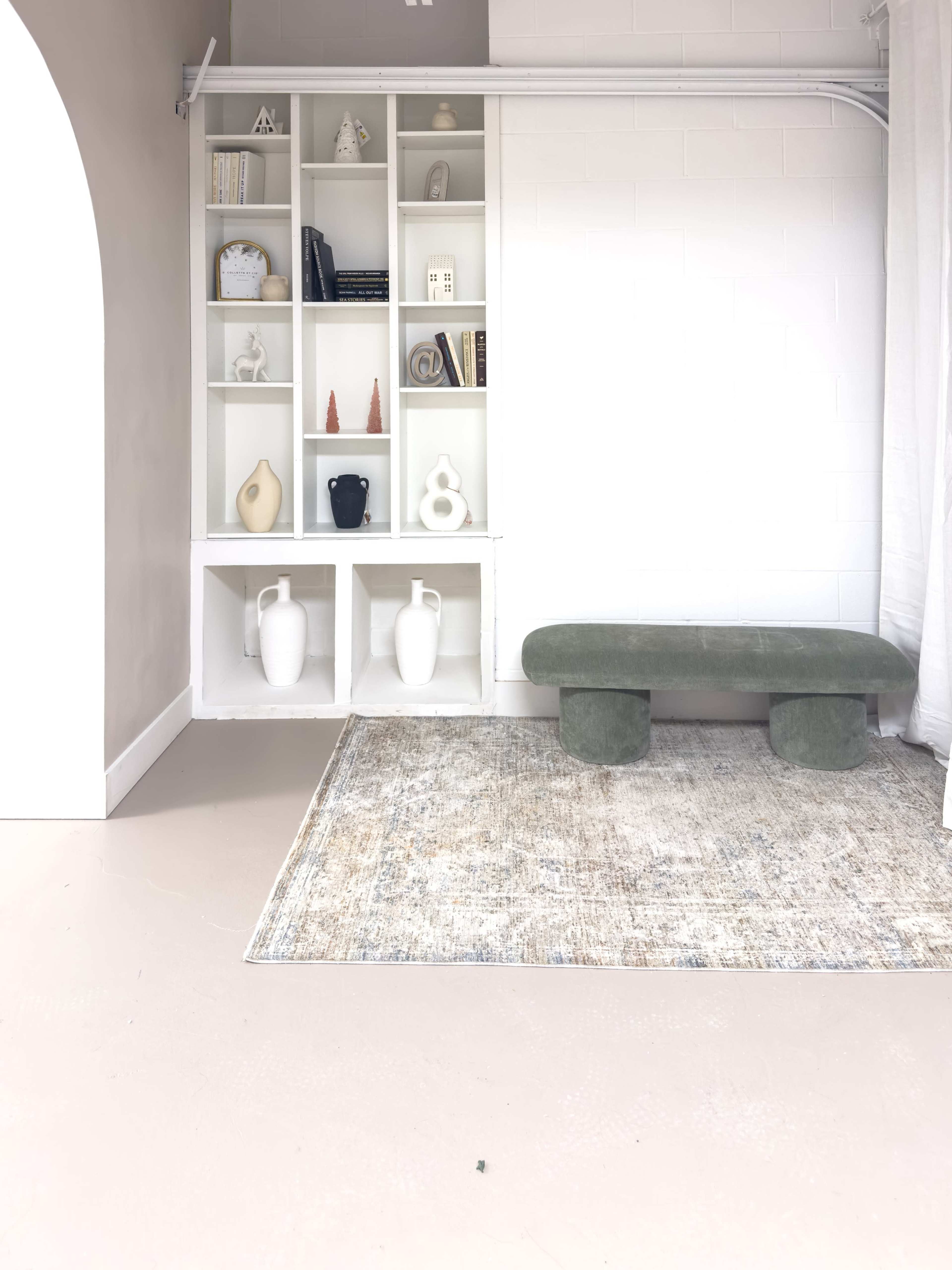 The image shows a minimalist interior featuring a white shelving unit holding decorative objects, a green bench, and a patterned rug on a light-colored floor.