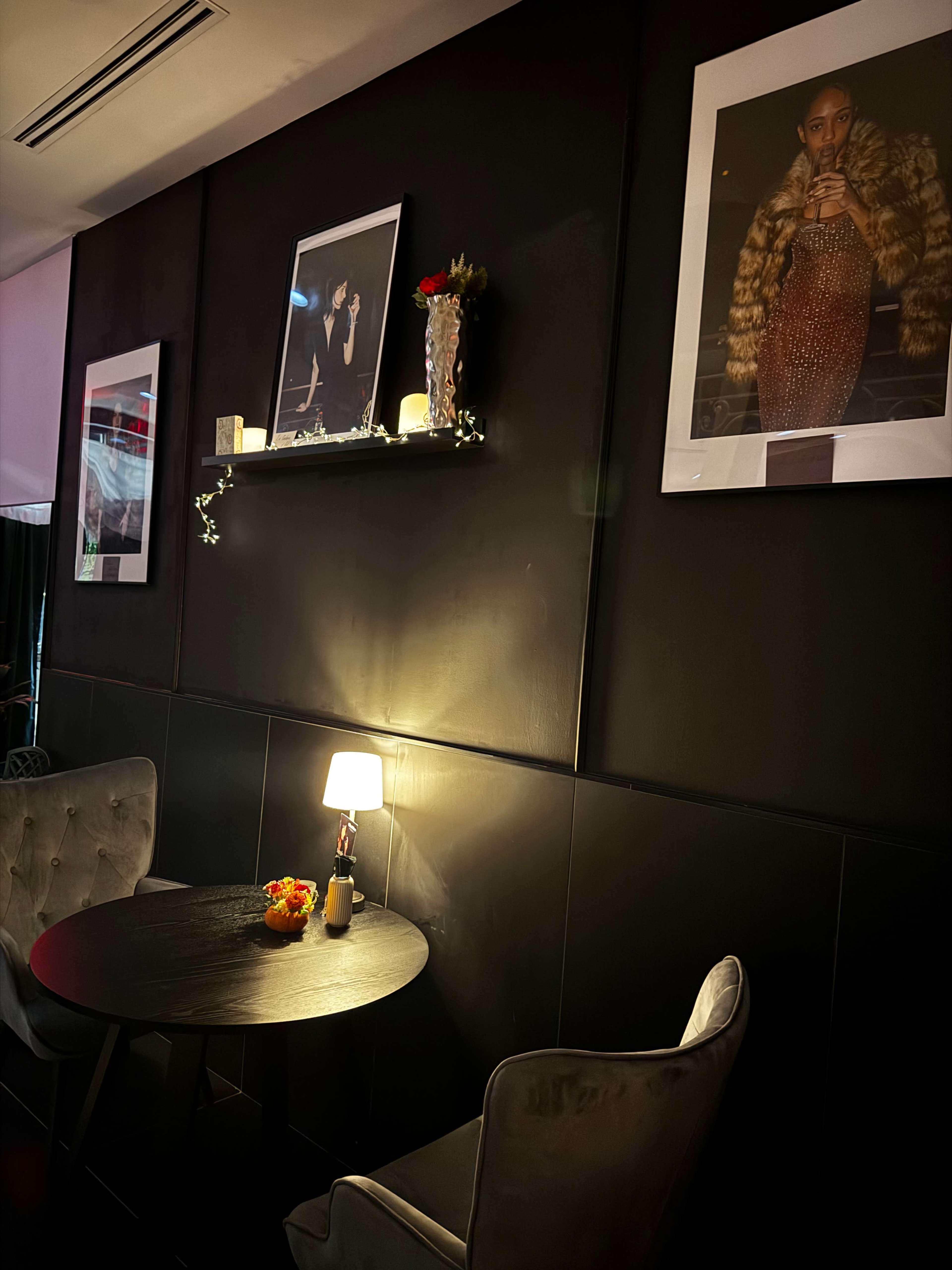 The image shows a dimly lit corner of a café or restaurant with a small round table, two plush chairs, and framed photographs on the dark wall.
