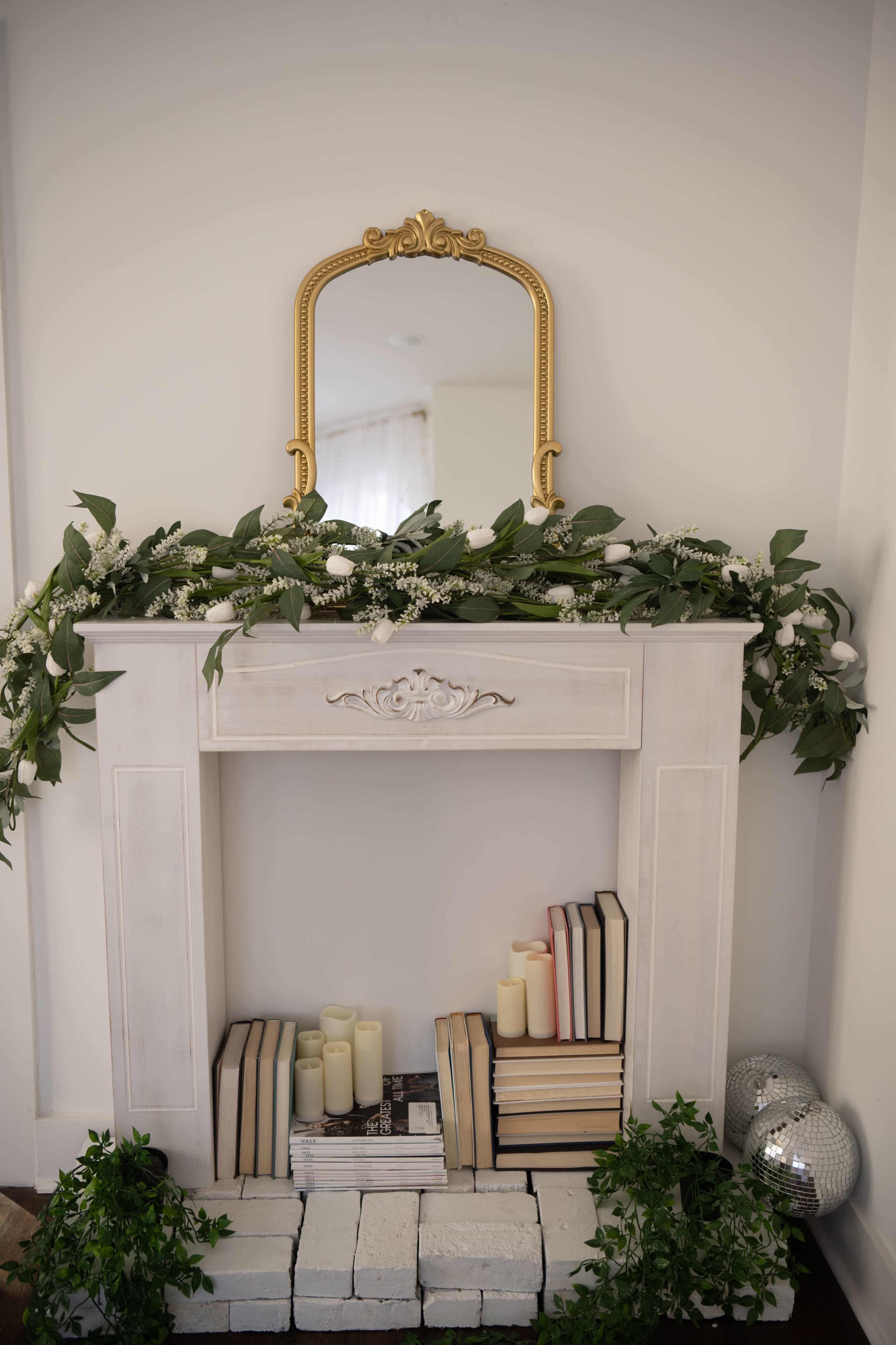 A white mantel is adorned with a gilded mirror, greenery, and stacks of books, while soft candles rest on a base of white bricks.
