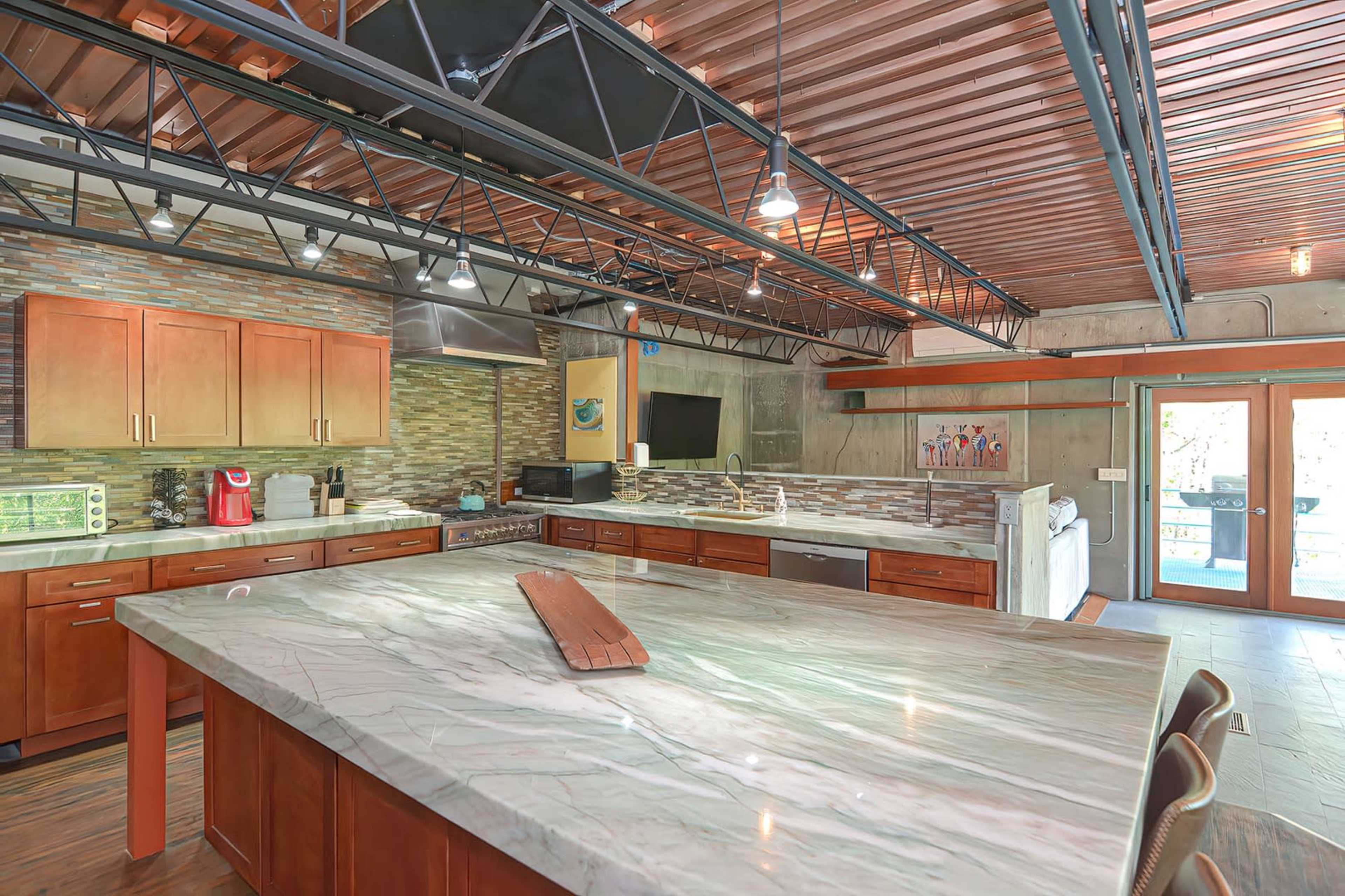 The kitchen features modern cabinets, a large marble island, and a stainless-steel appliance setup under a metal beam ceiling.