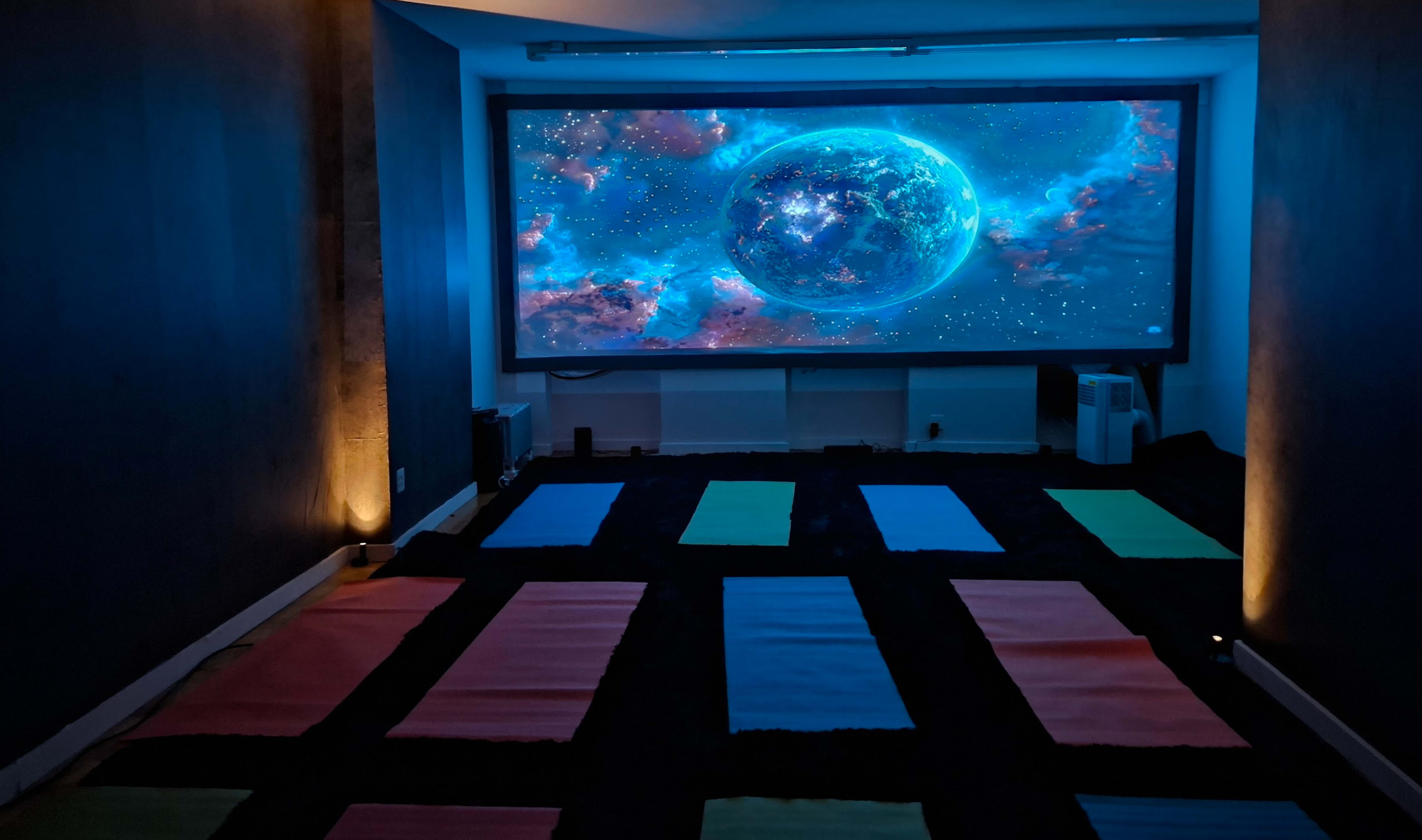 The image shows a dimly lit room with a large projection screen displaying a cosmic scene, surrounded by colorful yoga mats arranged on the floor.