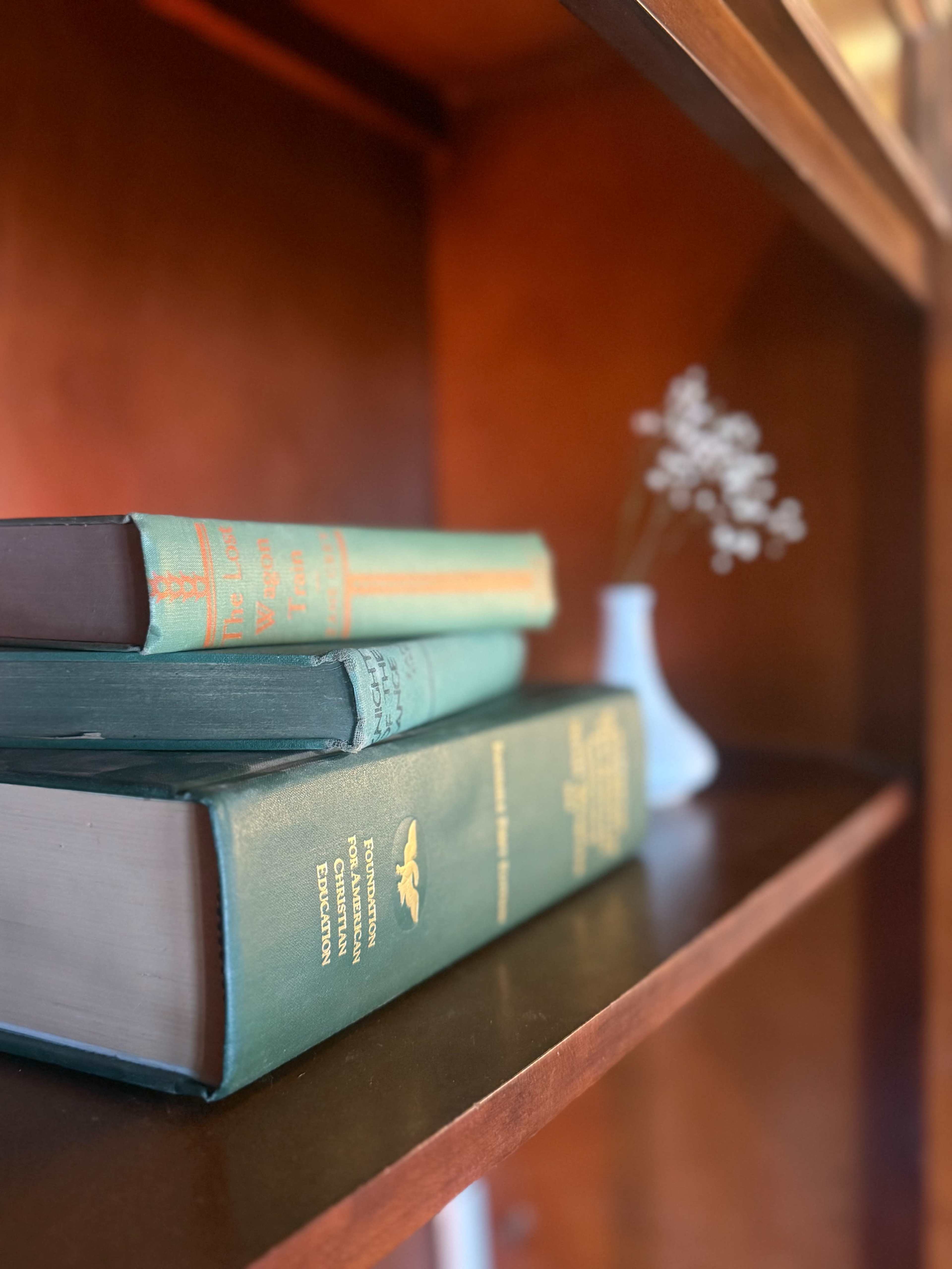 A stack of three hardcover books sits on a wooden shelf next to a small vase with delicate white flowers.