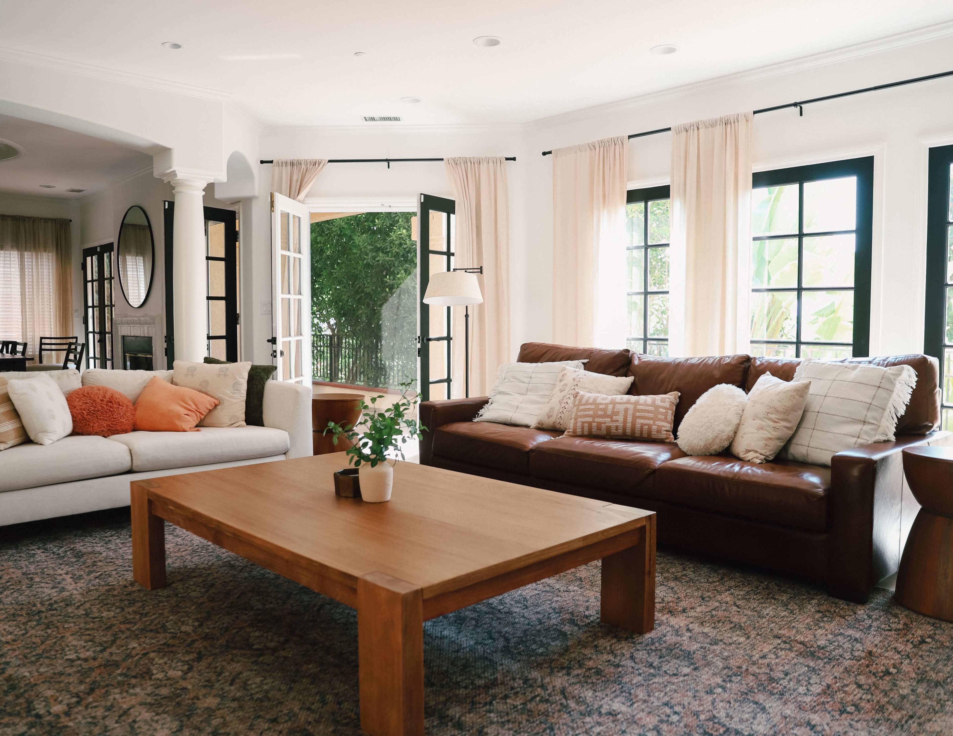 A living room features a brown leather sofa and a light-colored couch, with a wooden coffee table at the center and large windows letting in natural light.