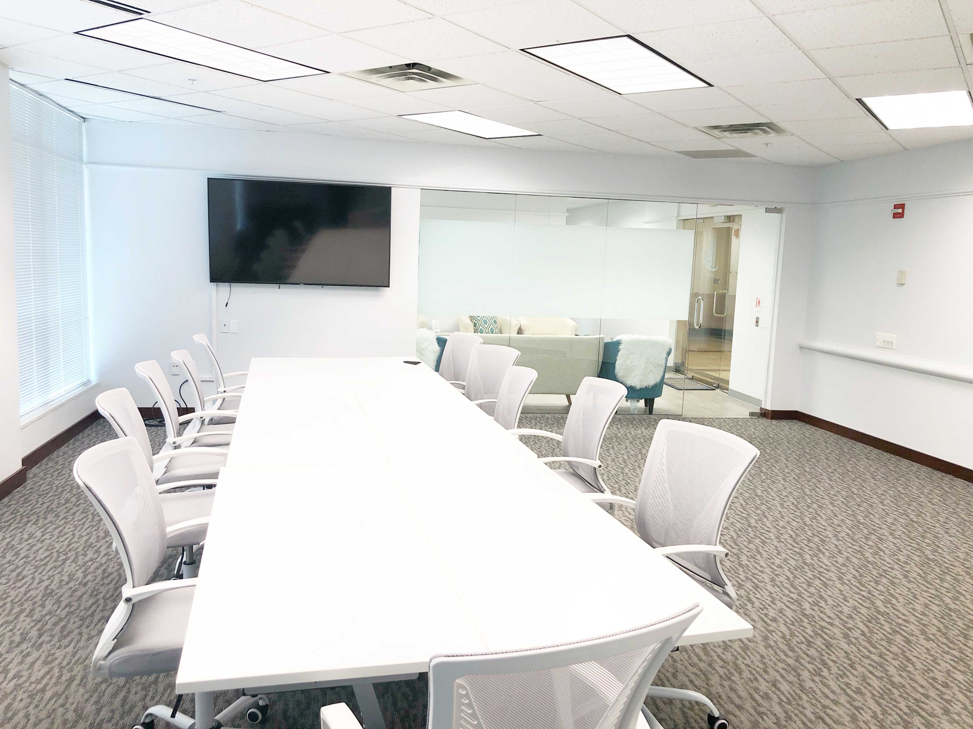 A modern conference room features a long white table surrounded by ergonomic chairs, with a large screen mounted on the wall and a glass partition revealing a separate lounge area.