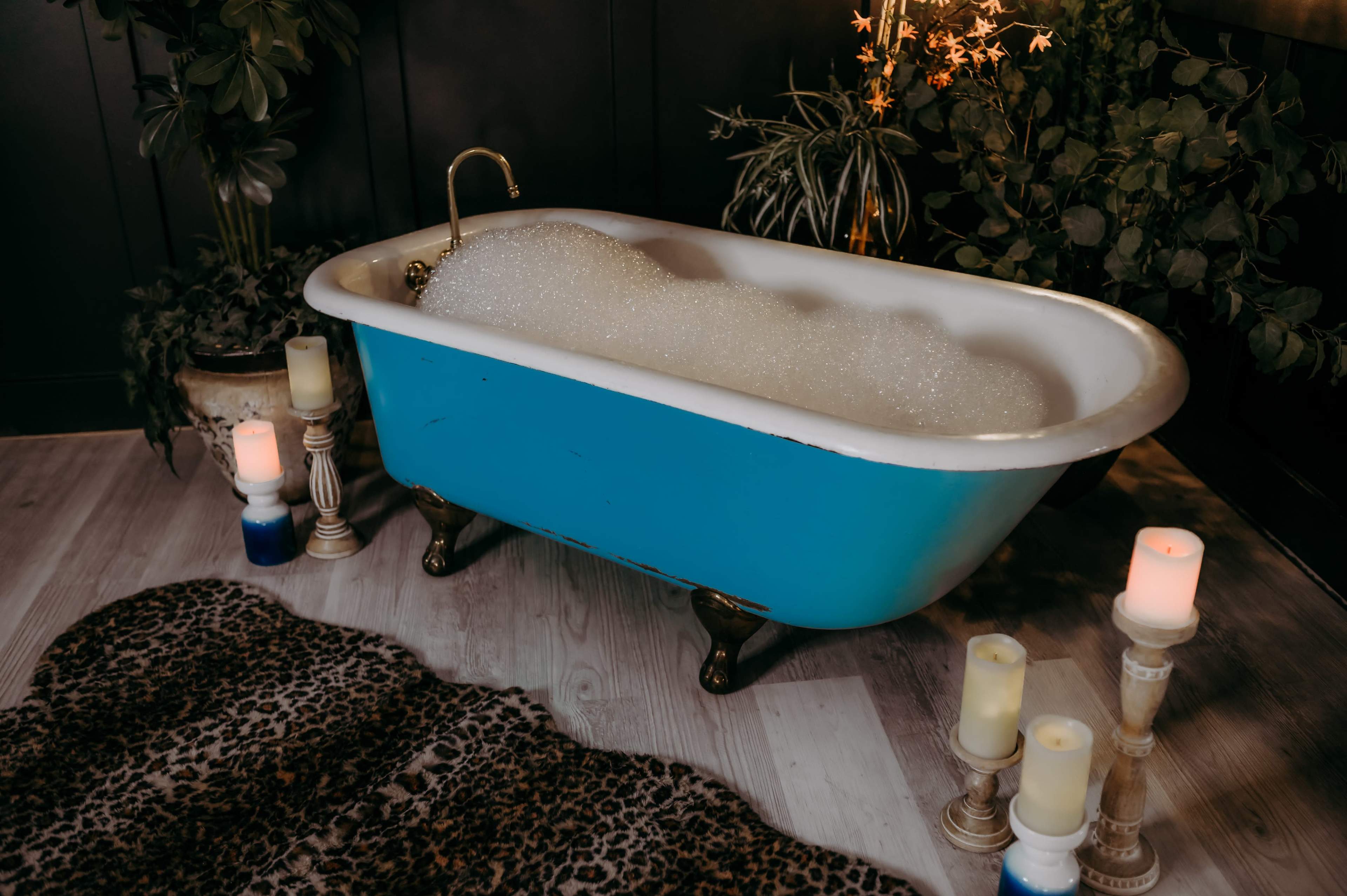 A vintage clawfoot bathtub filled with bubbles sits on a wooden floor, surrounded by potted plants and candles.