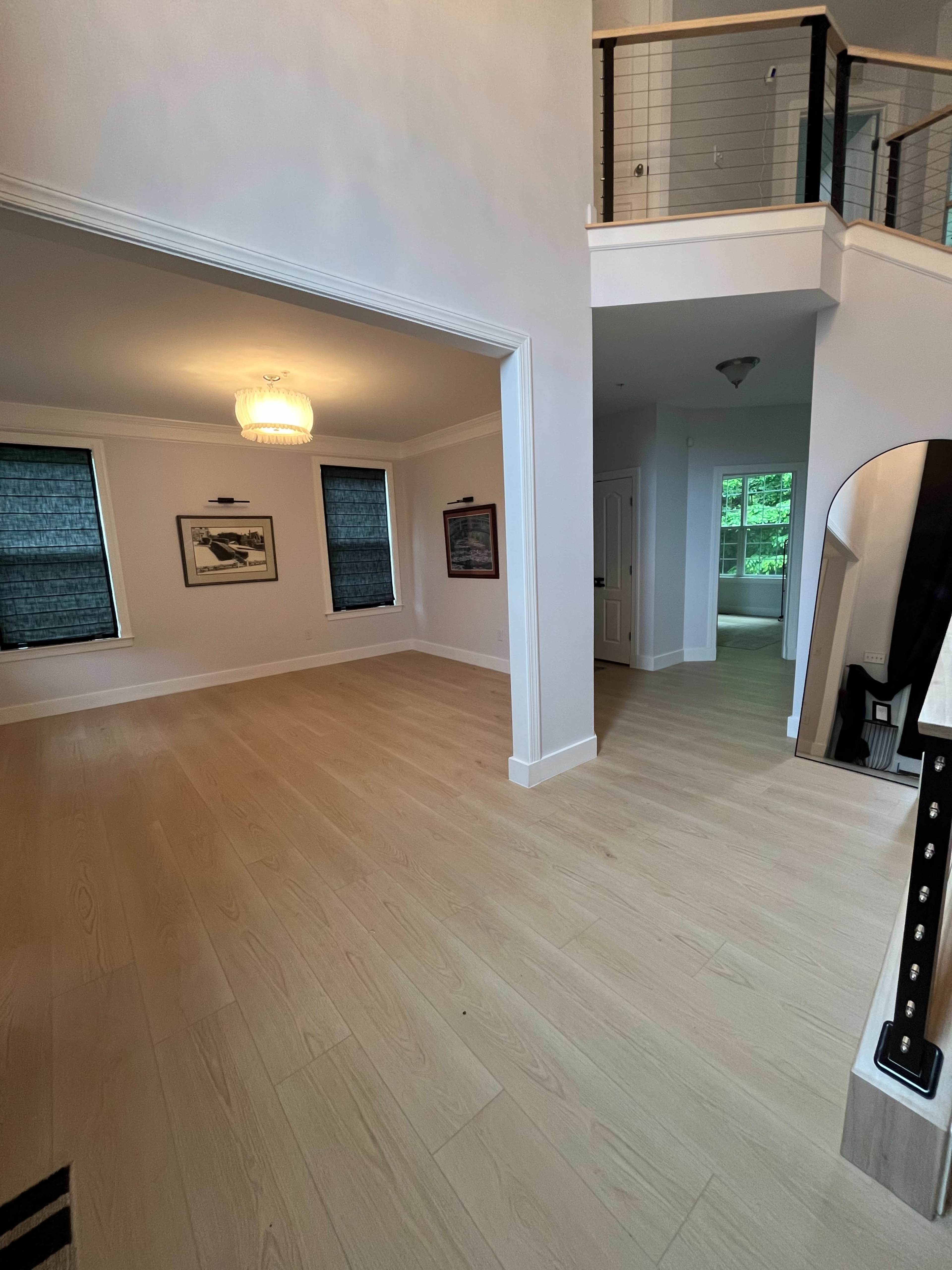The image shows a spacious room with light wood flooring, two framed pictures on the wall, and large windows with dark shades.