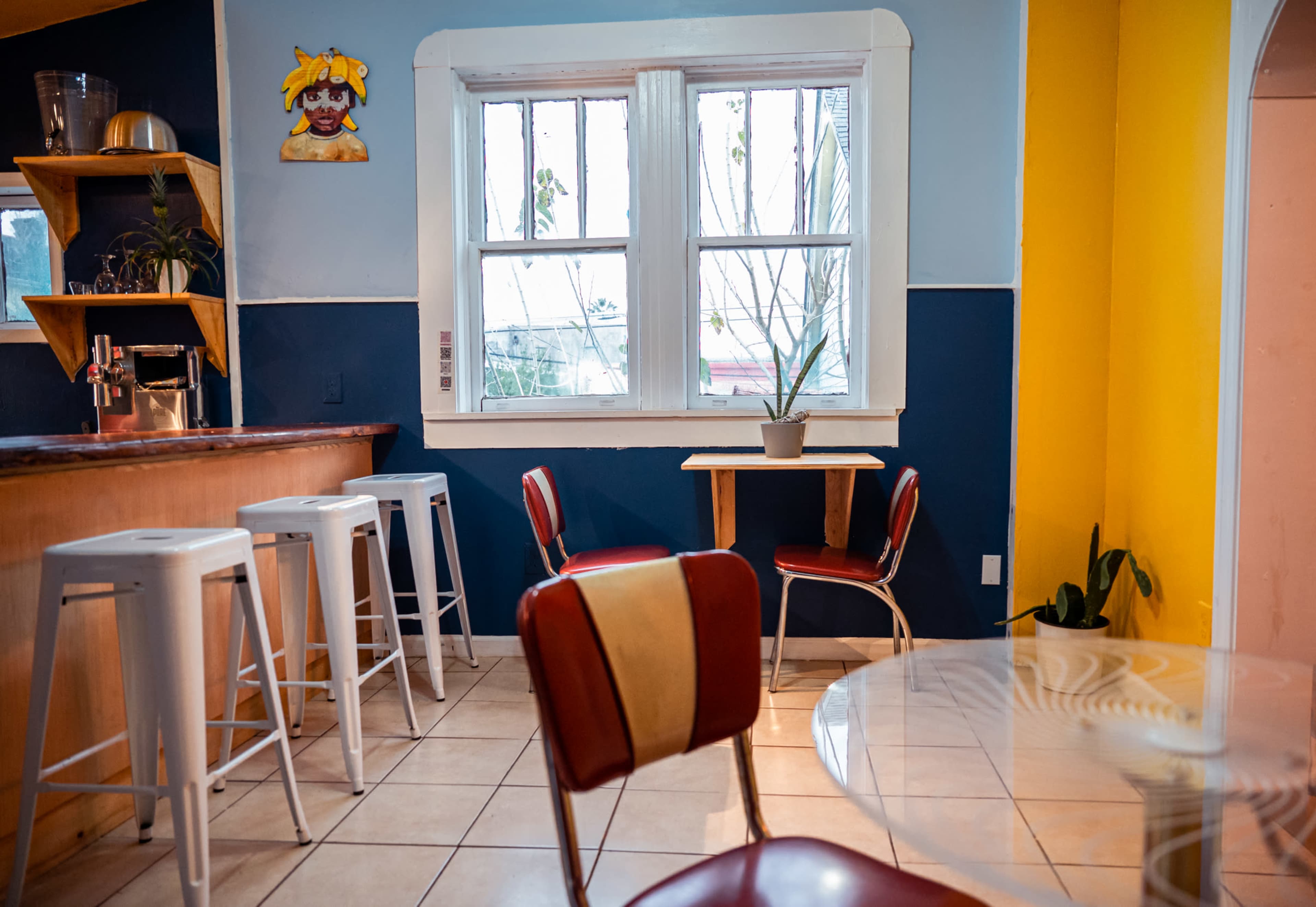 A cozy café interior with a bar counter, several stools, and a small table surrounded by red chairs near a window.