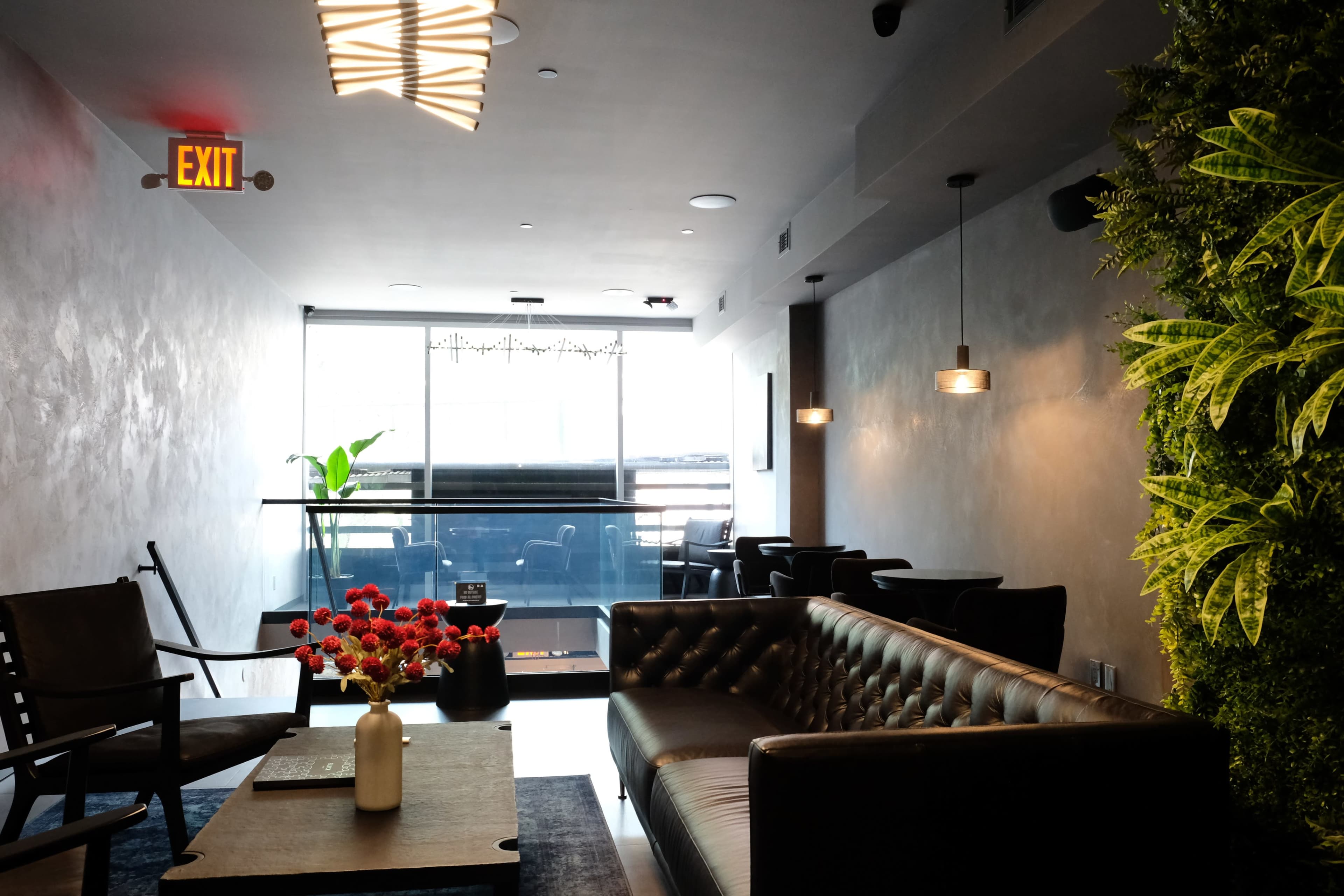 The image shows a modern lounge area featuring a brown leather sofa, a wooden table with flowers, and a large window with natural light.