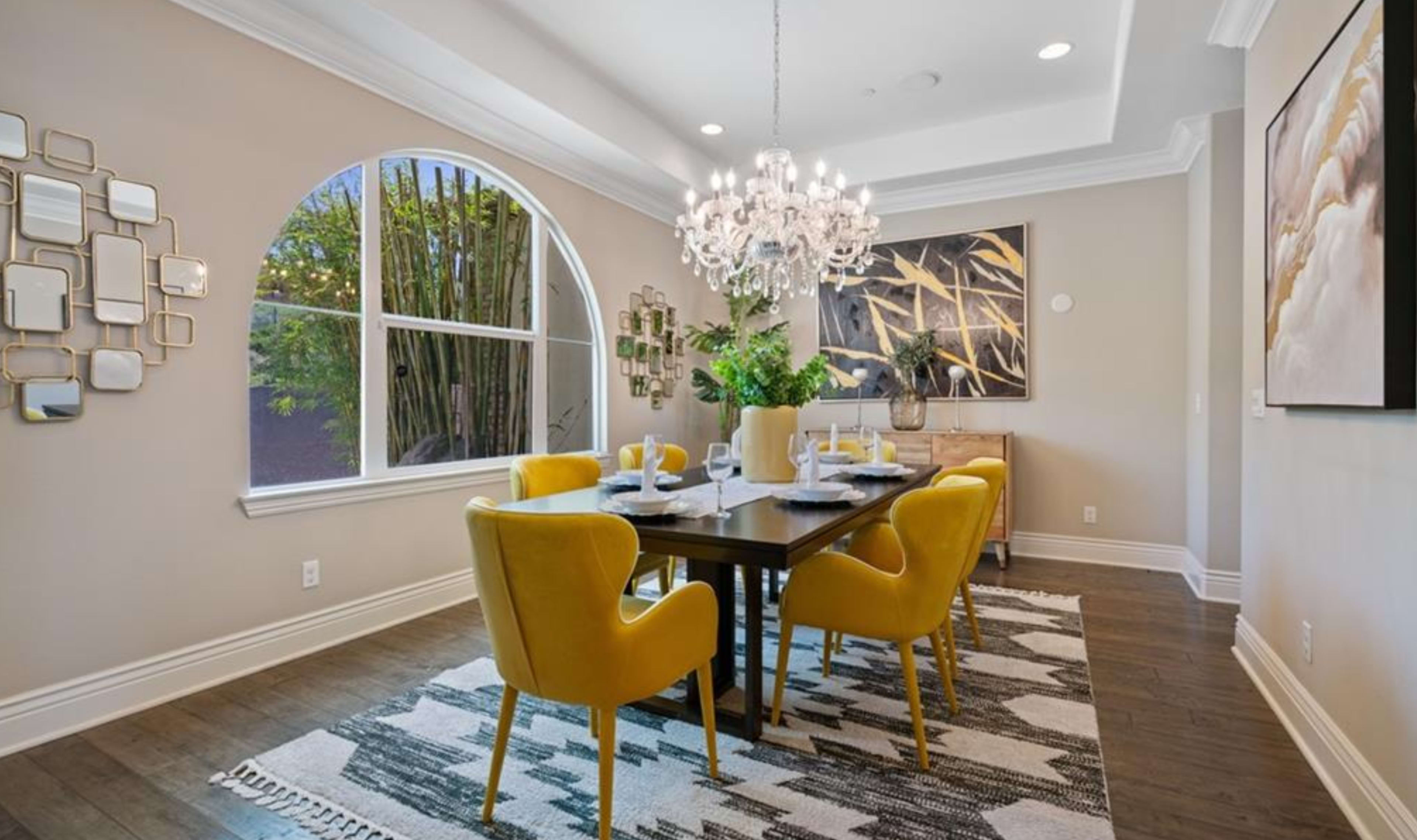 A modern dining room features a dark wooden table surrounded by yellow chairs, with a large chandelier overhead and an arched window letting in natural light.