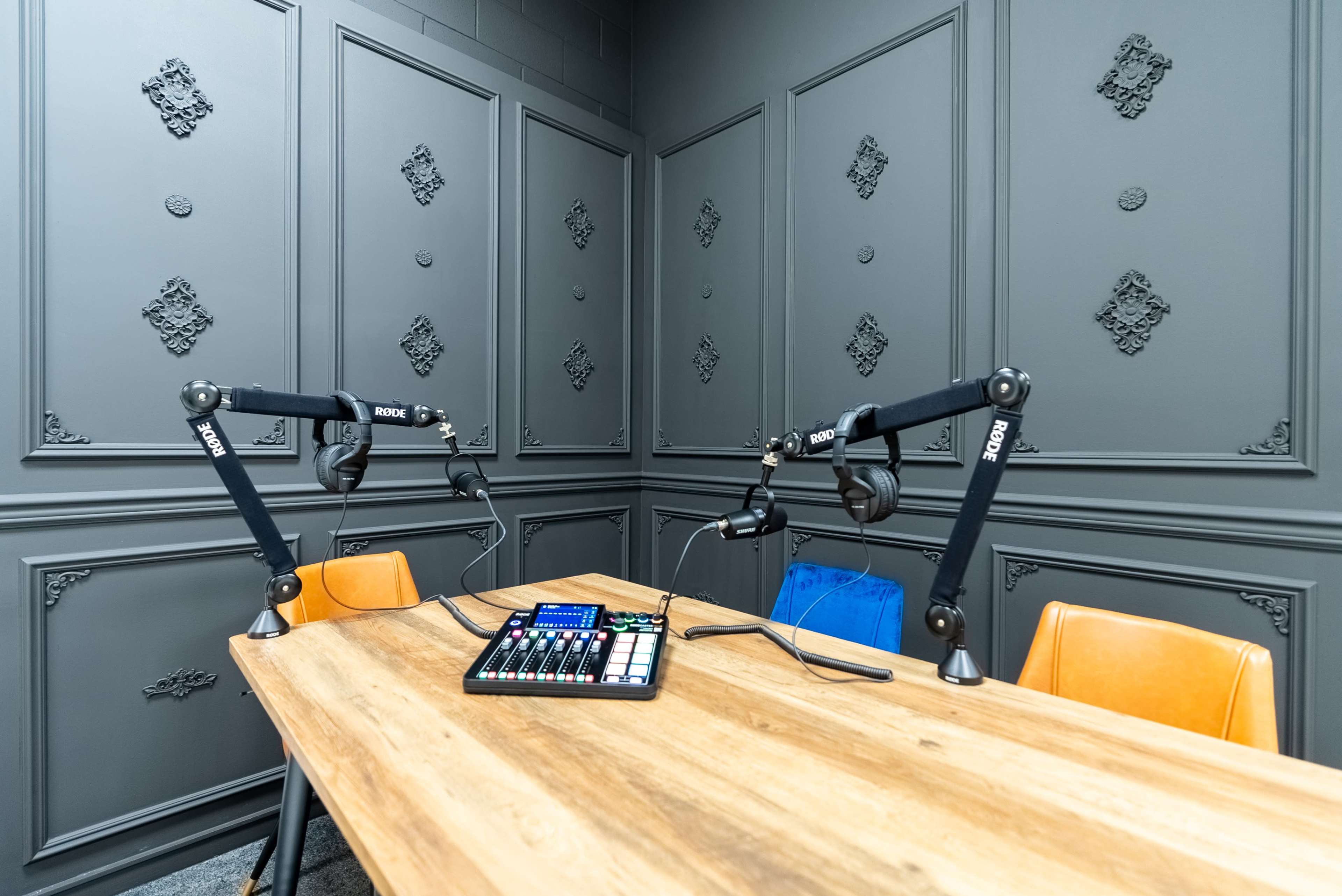 A modern recording studio setup with two microphone arms and a control panel on a wooden table against a textured gray wall.