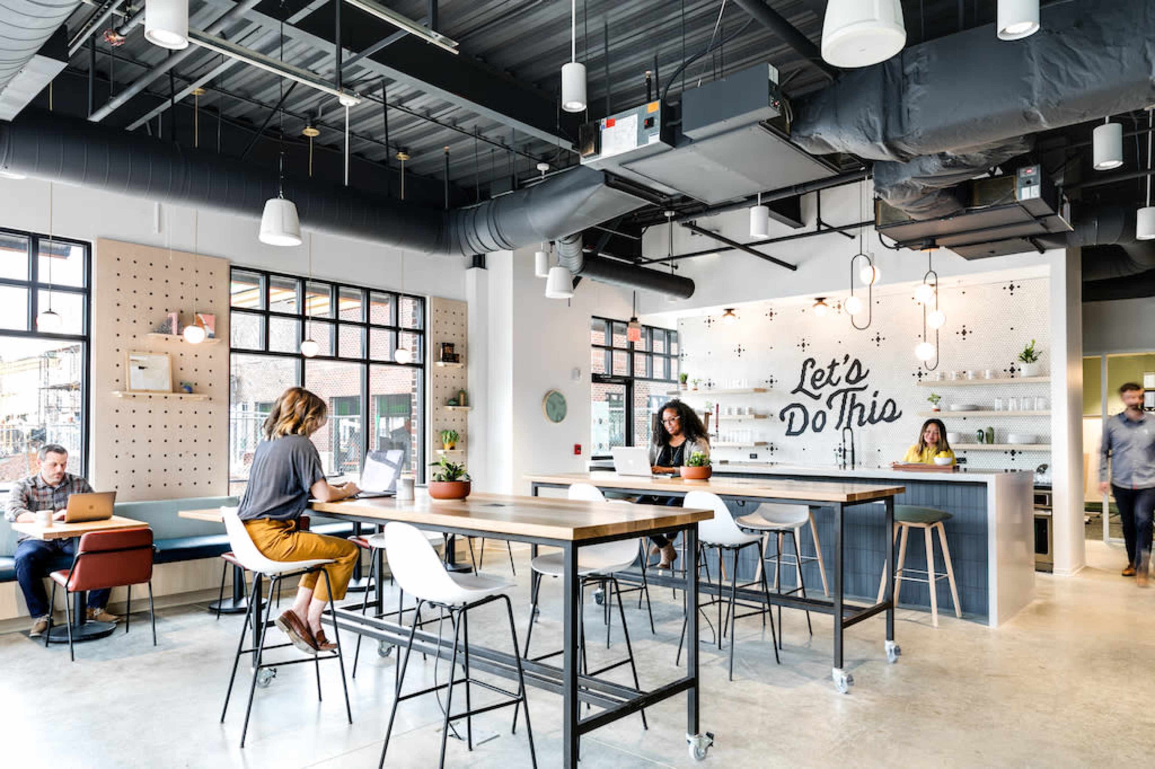 A modern co-working space with people working at tables, a stylish bar area, and large windows providing natural light.