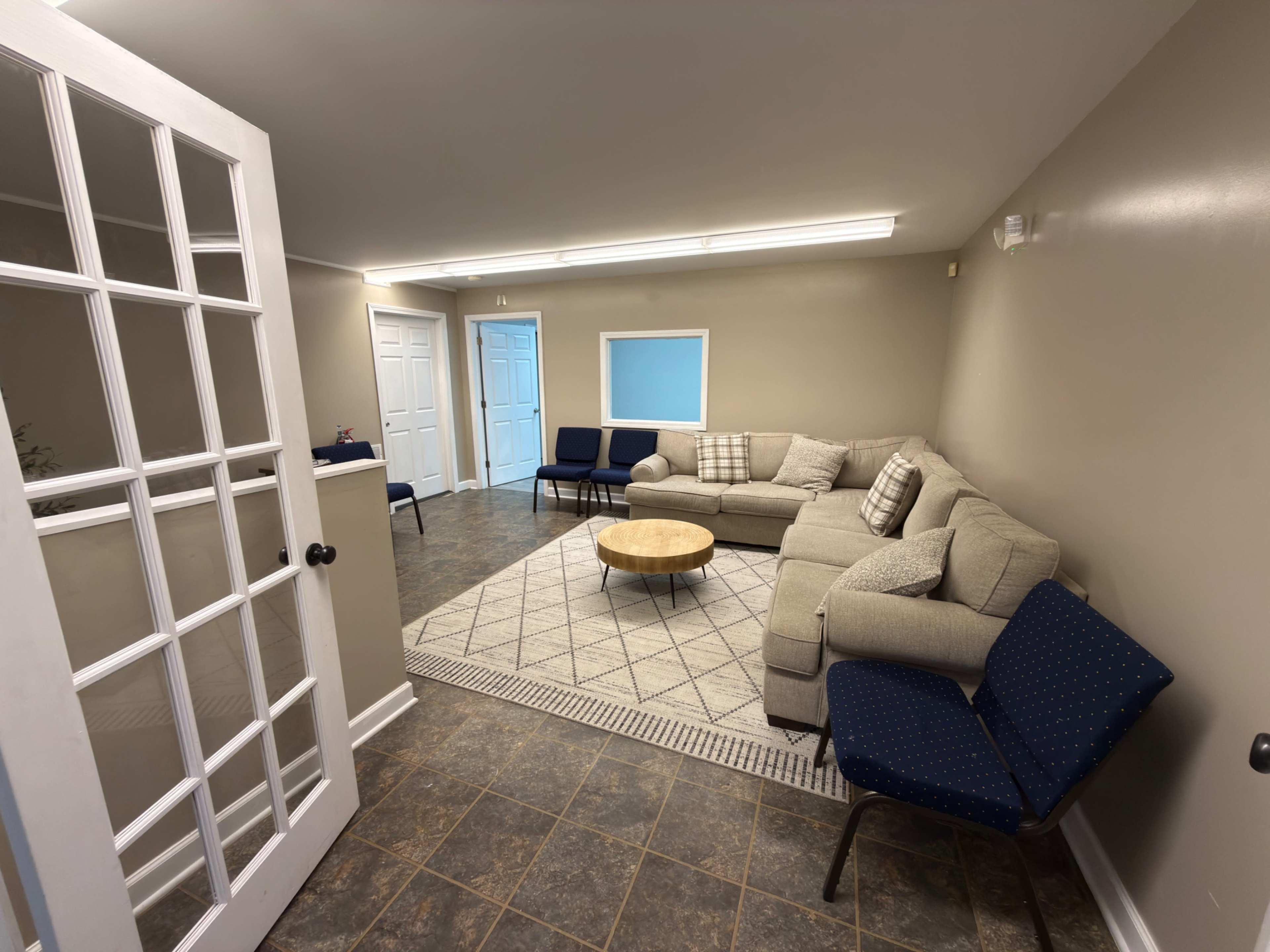 The image shows a neatly arranged room with a sectional couch, a round coffee table, and several chairs against the walls, all set on a patterned rug.
