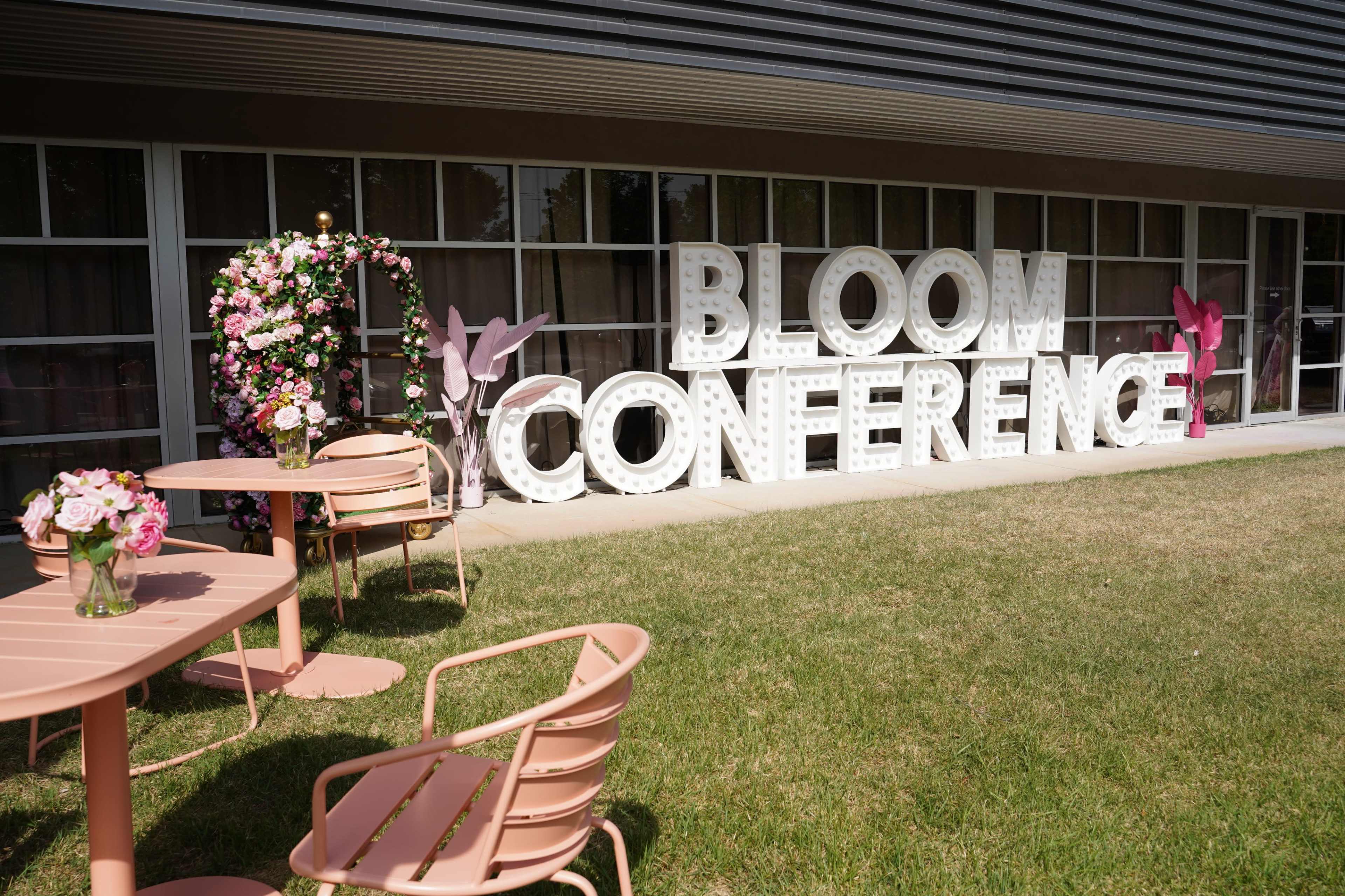 The image shows a garden area with a large sign reading "BLOOM CONFERENCE" alongside floral decorations and pink outdoor furniture.