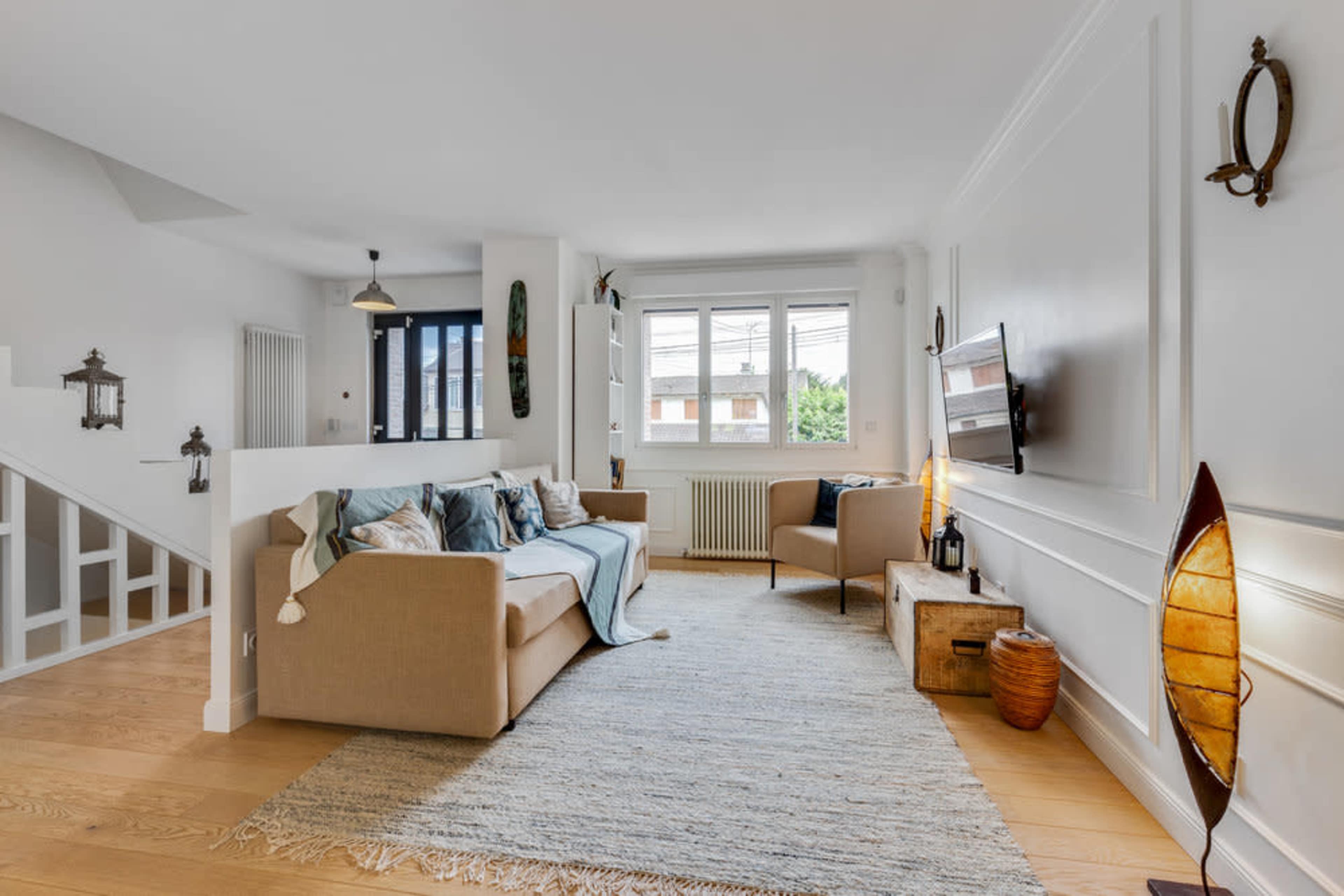 A light and airy living room features a beige sofa, a wooden armchair, and a decorative rug on a hardwood floor.