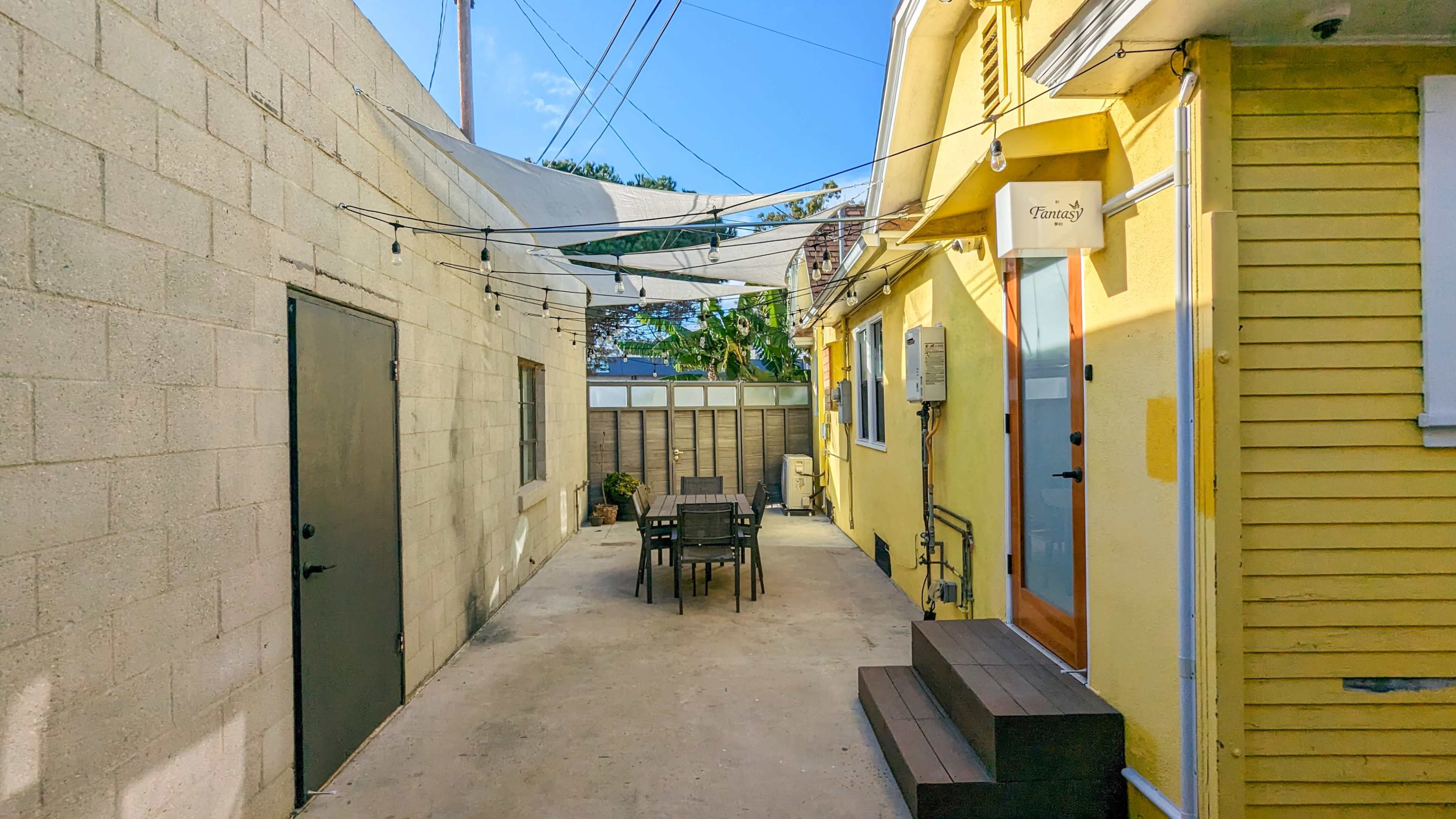 A narrow outdoor courtyard between two buildings, featuring a table and chairs, string lights overhead, and a bright yellow wall on one side.
