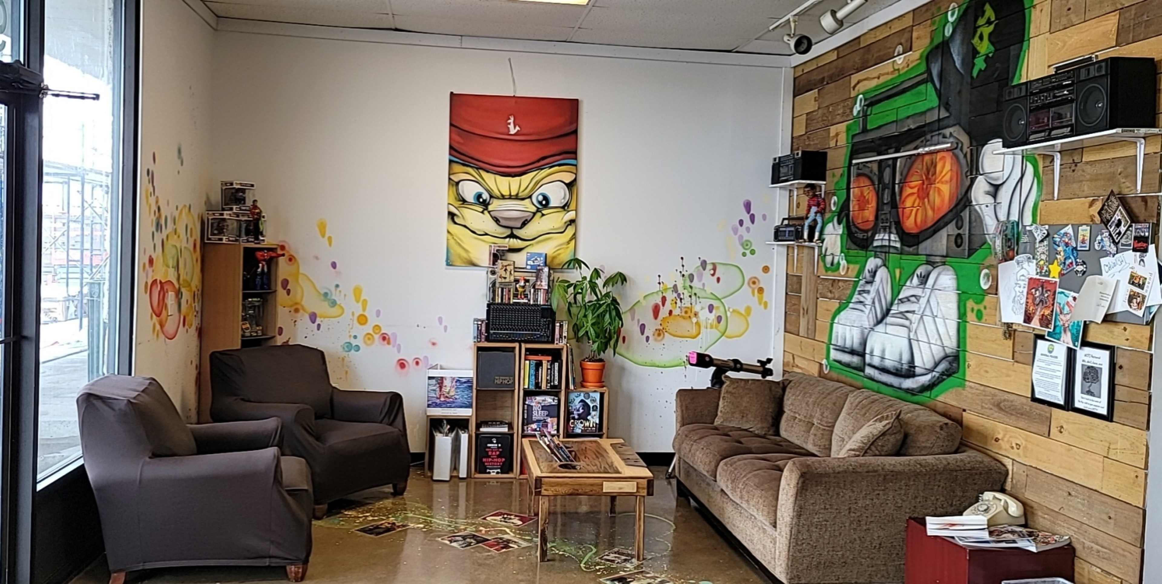 A small lounge area with two gray chairs, a brown couch, a wooden coffee table, and colorful graffiti on the walls, including a large painted character and musical equipment.