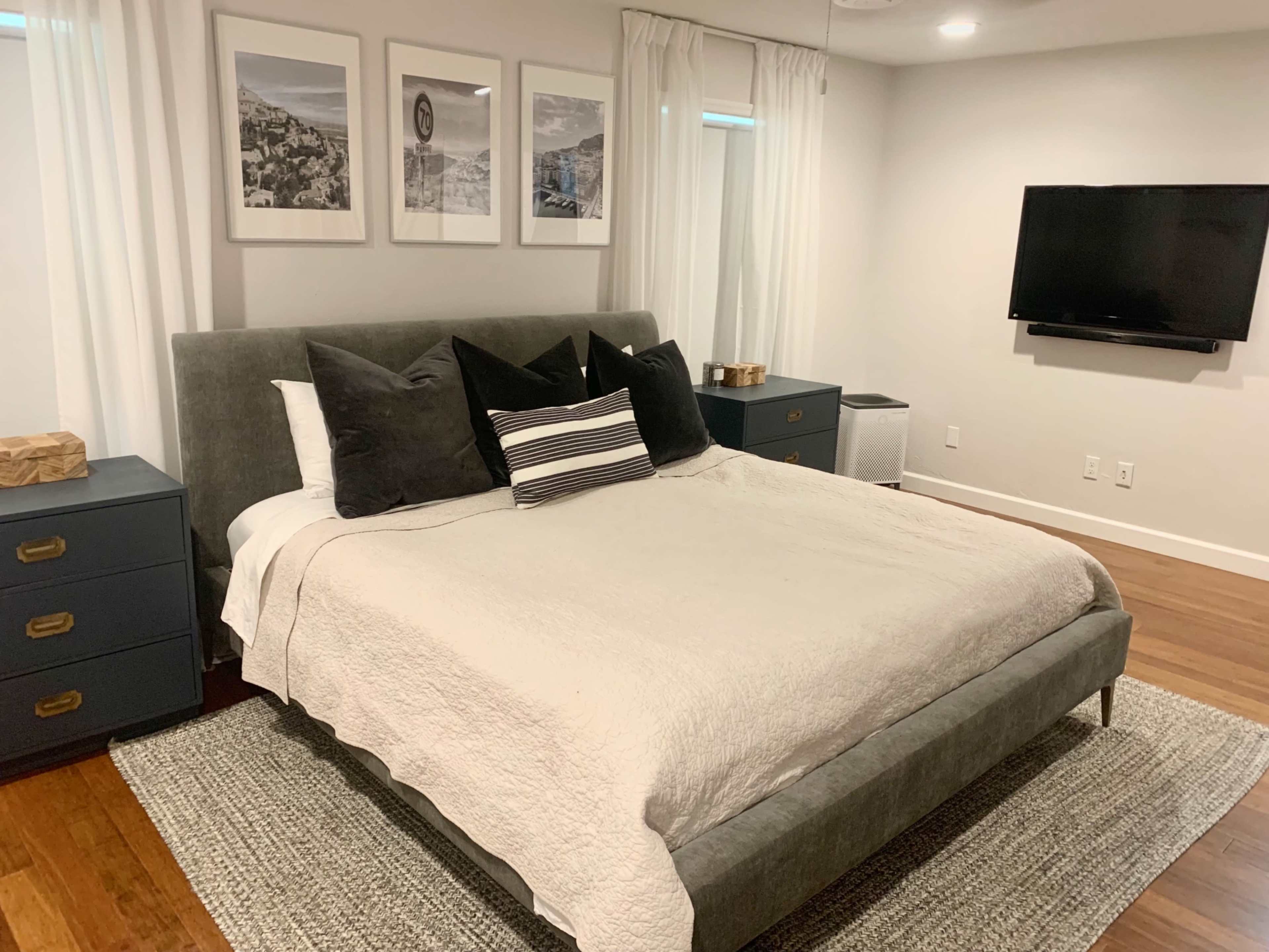 The image depicts a neatly arranged bedroom with a gray upholstered bed, two dark blue nightstands, and a wall-mounted television.