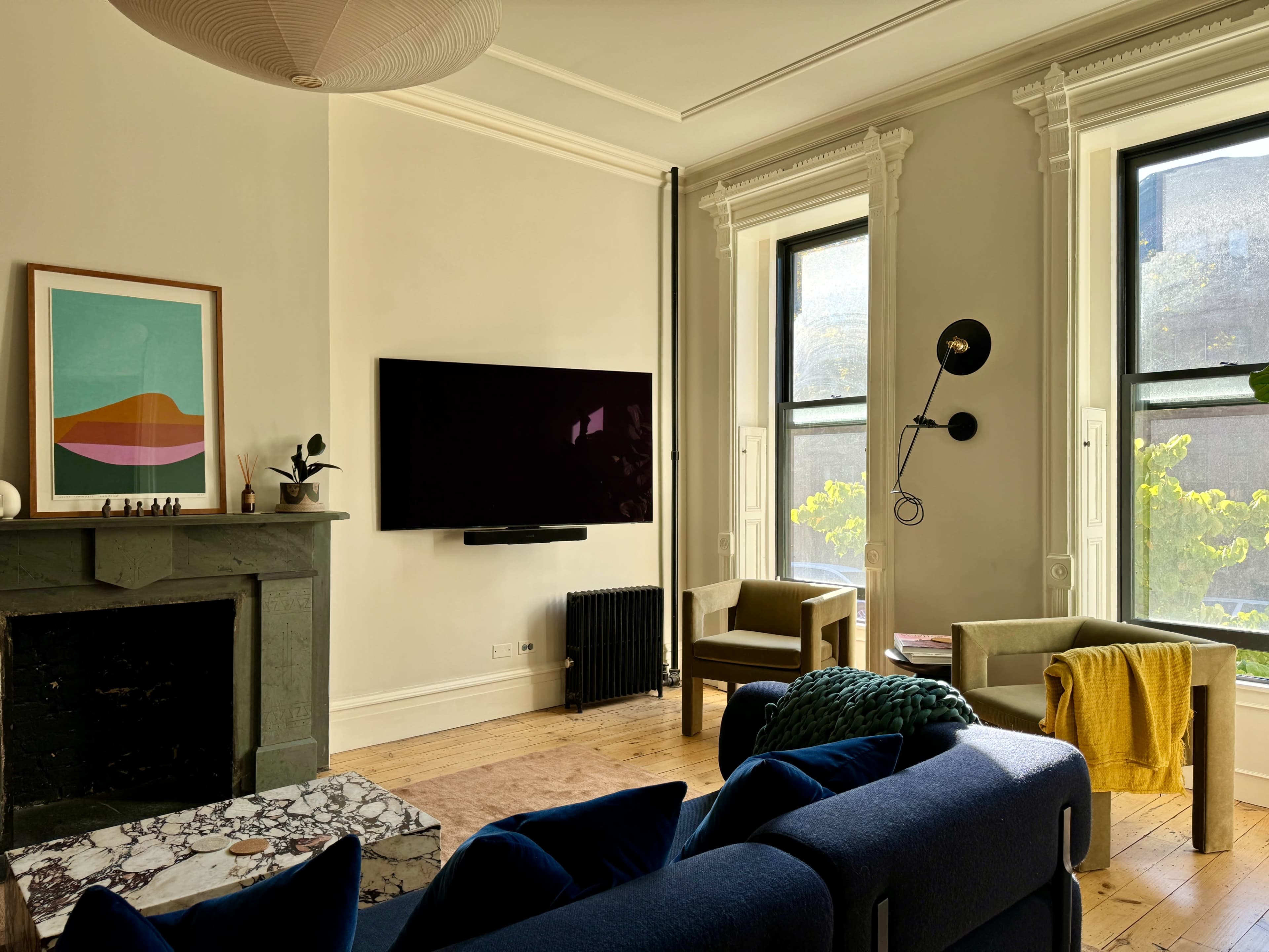The living room features a dark blue sofa facing a large television mounted above a marble fireplace, with two armchairs and a window providing natural light.