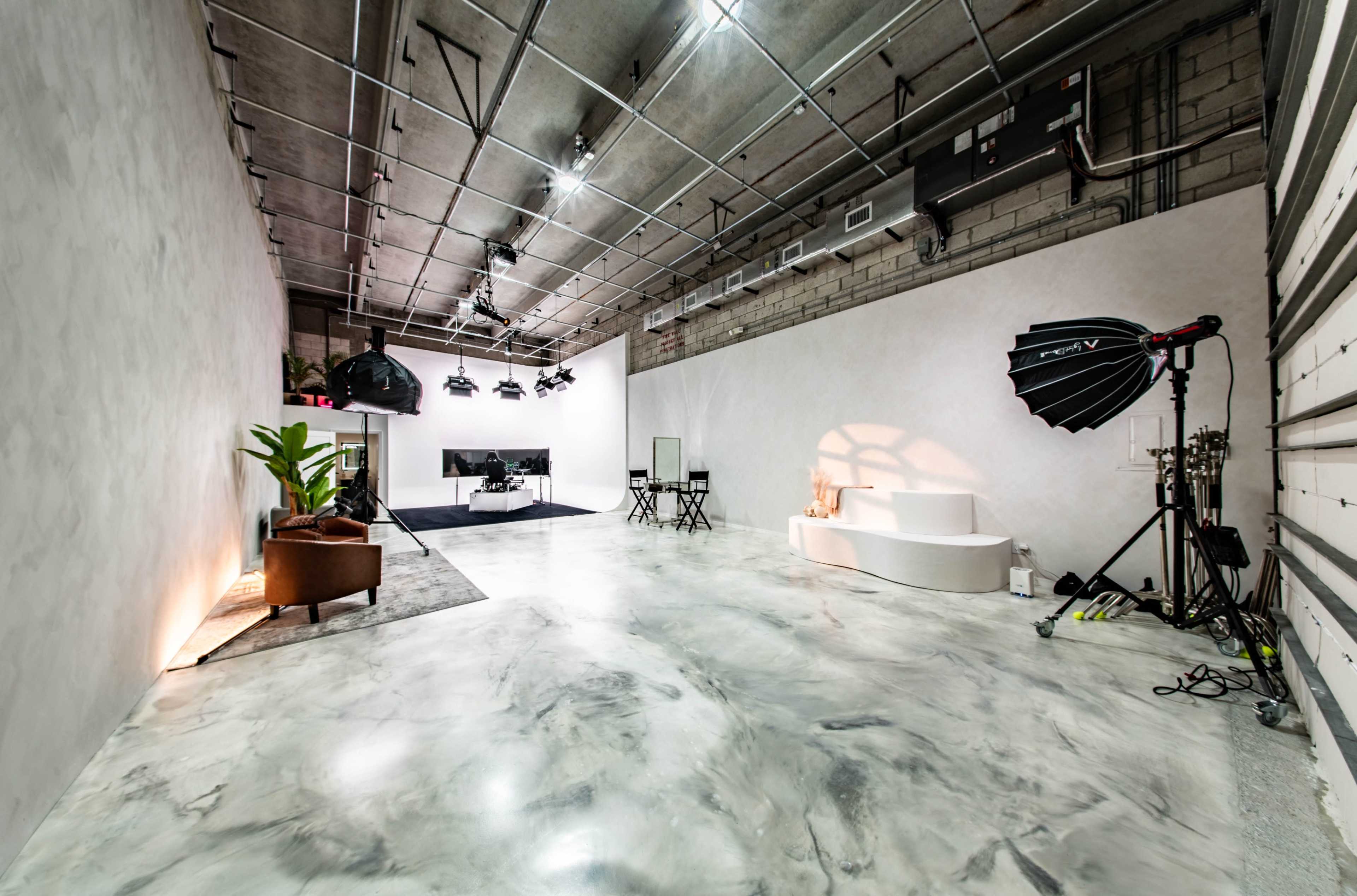 The image shows a spacious, minimalistic studio with polished concrete floors, soft lighting, and photography equipment arranged around the area.