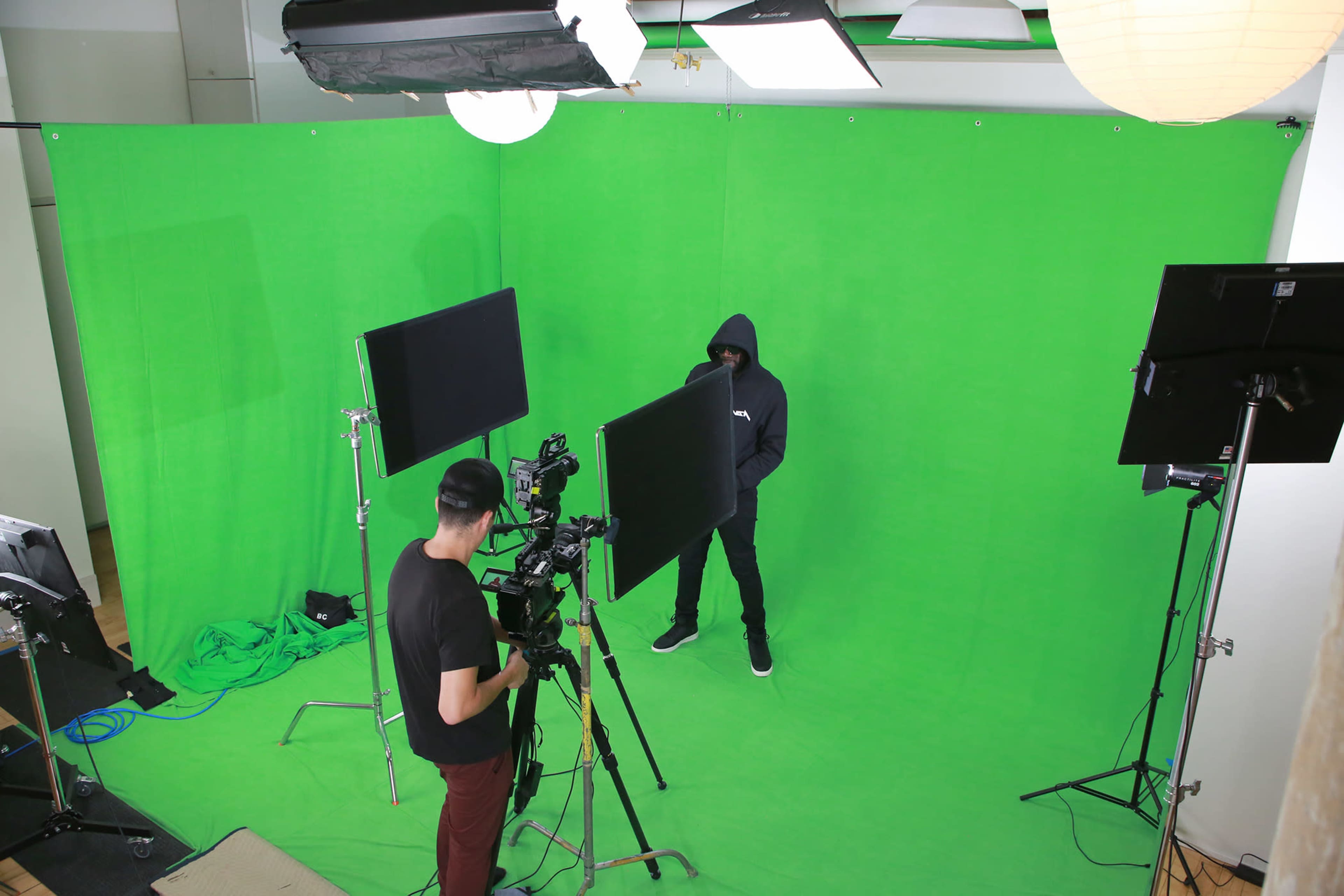 A person in a hoodie stands in front of a green screen while another person operates a camera nearby.