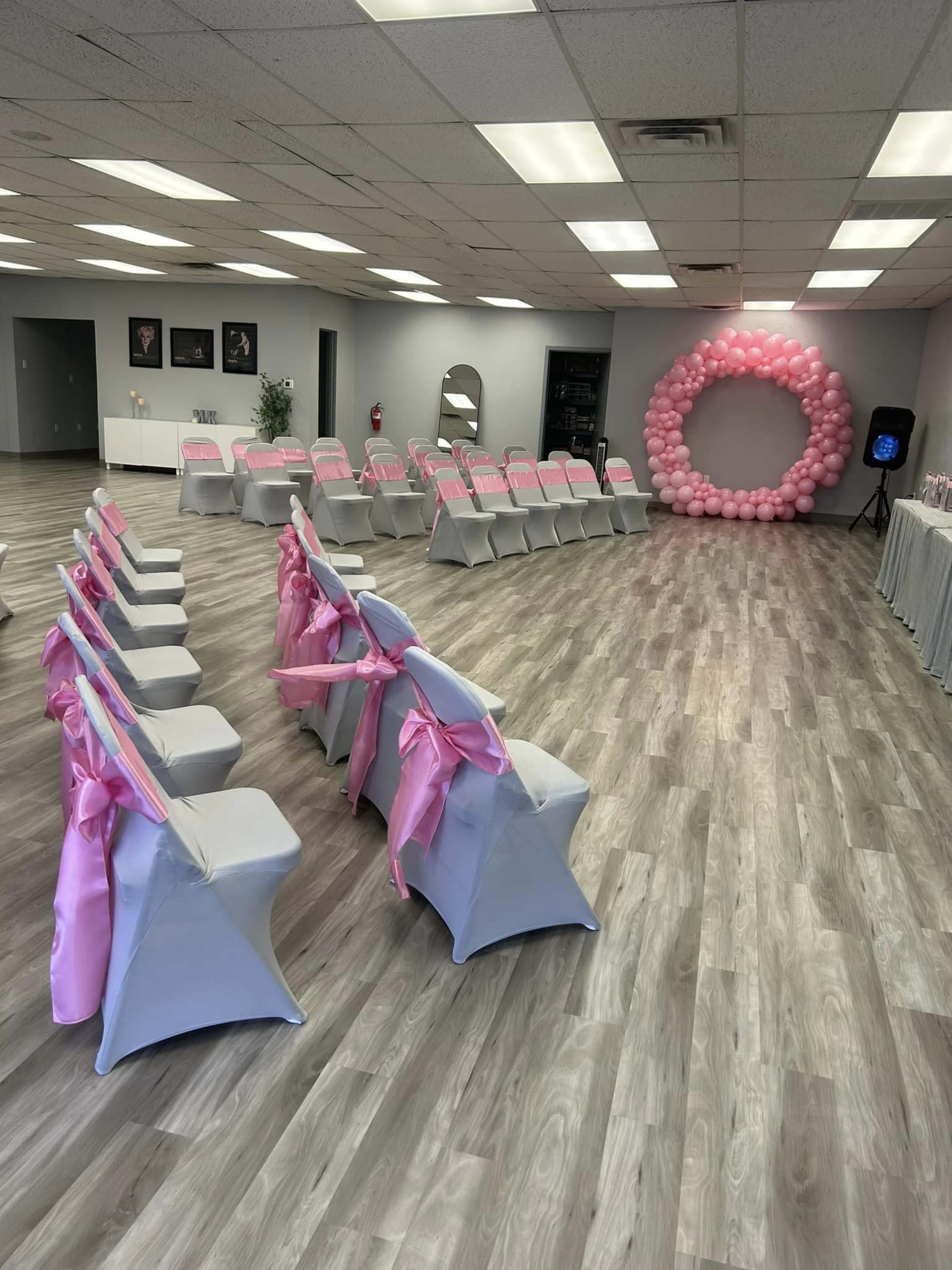The image shows an event space arranged with rows of chairs draped in gray covers and pink sashes, facing a round balloon arch at one end.