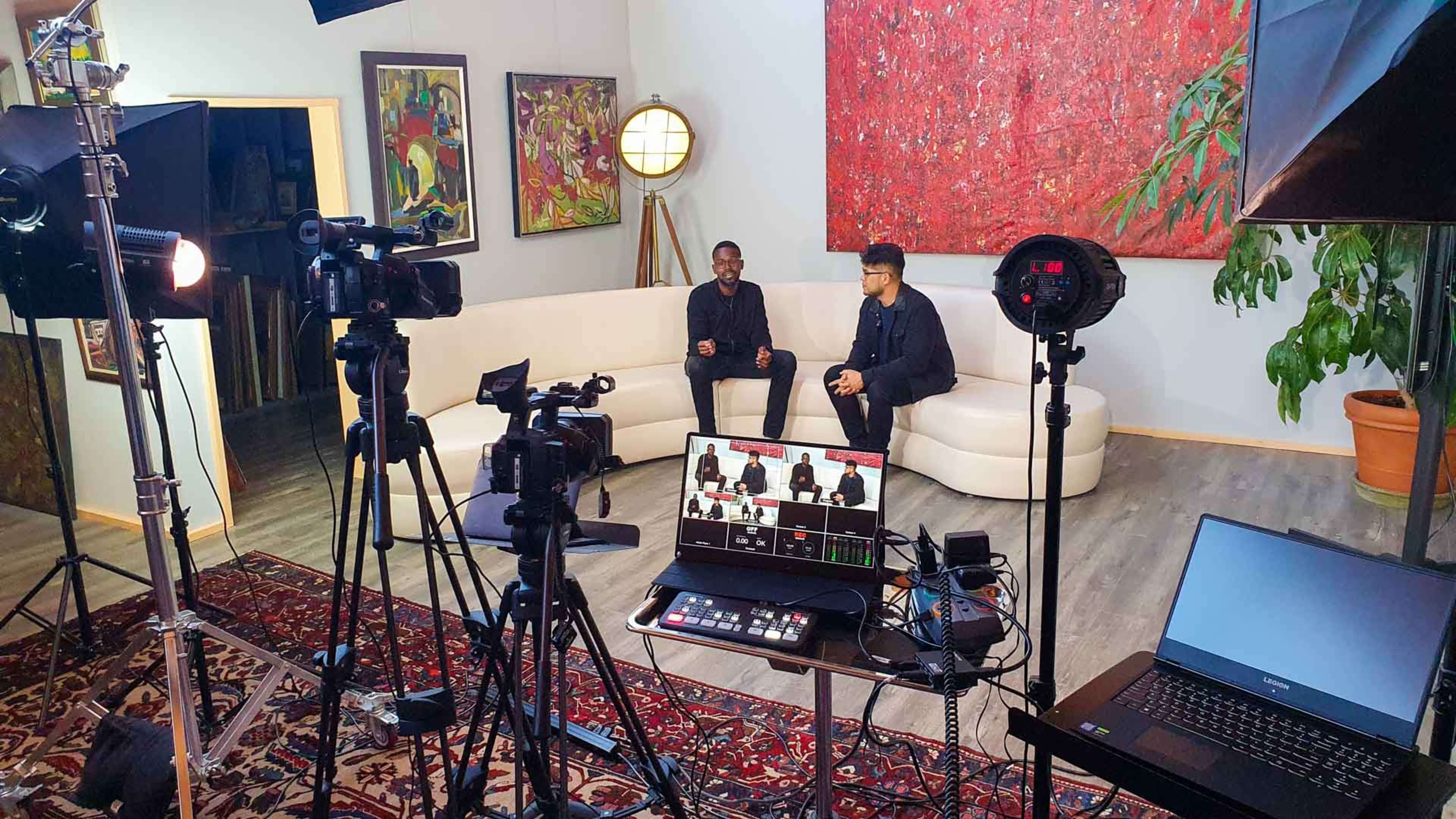 A well-equipped video production studio featuring two seated individuals on a white couch, surrounded by cameras, lighting equipment, and a control panel displaying a live feed.