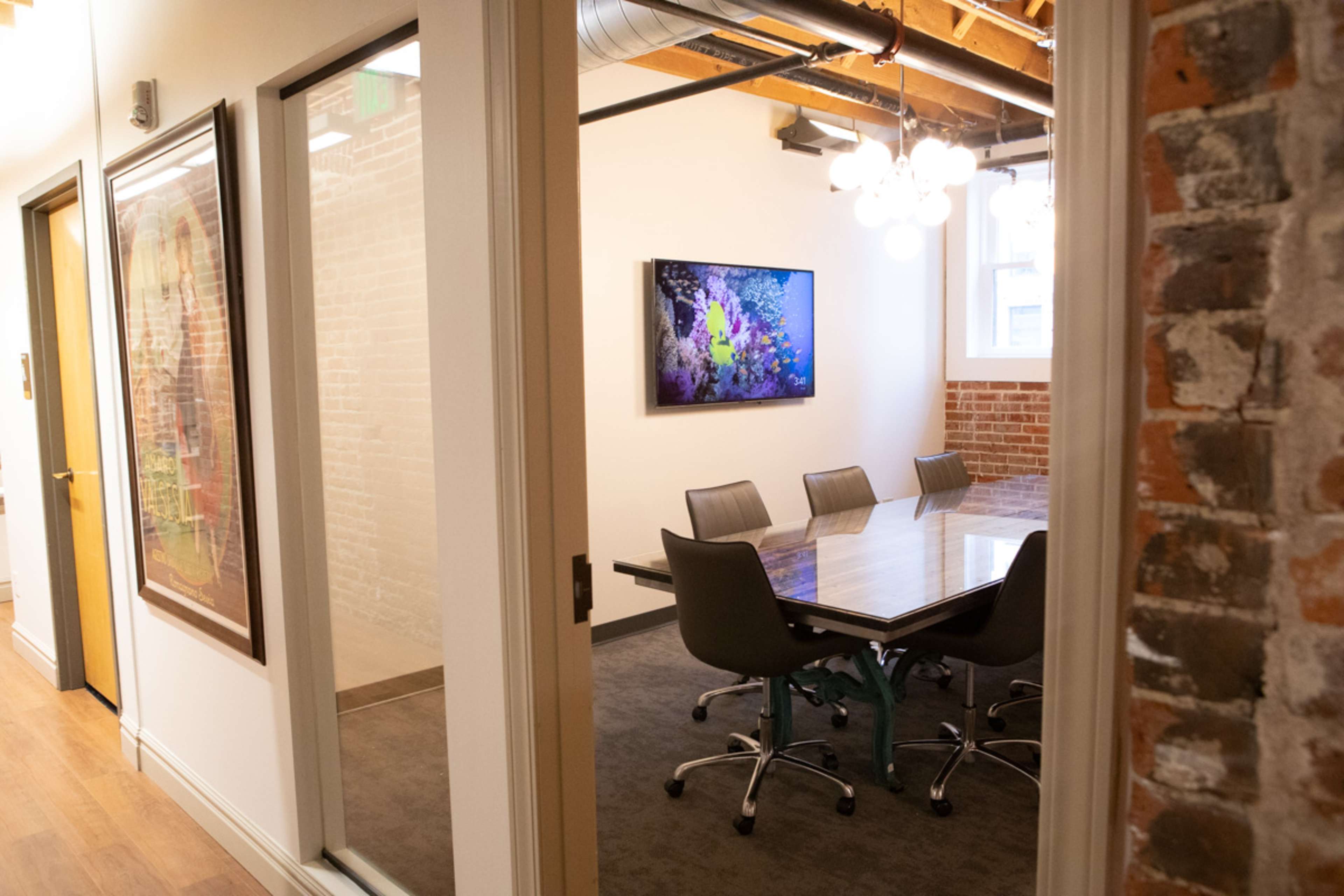 A conference room with a glass table and six chairs, illuminated by a wall-mounted screen and natural light from a nearby window.