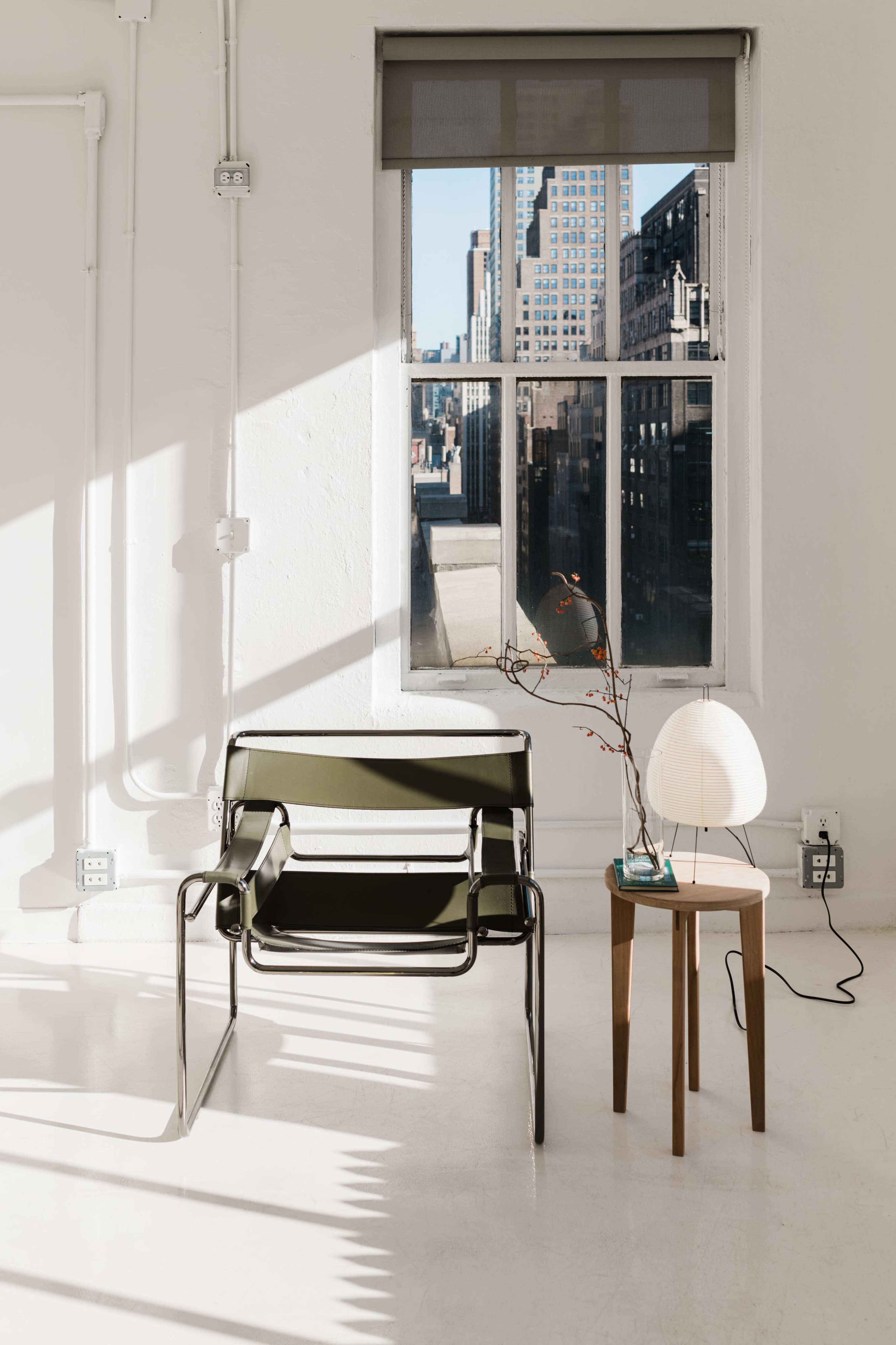 A green chair and a small wooden table with a lamp sit beside a large window showcasing a cityscape.