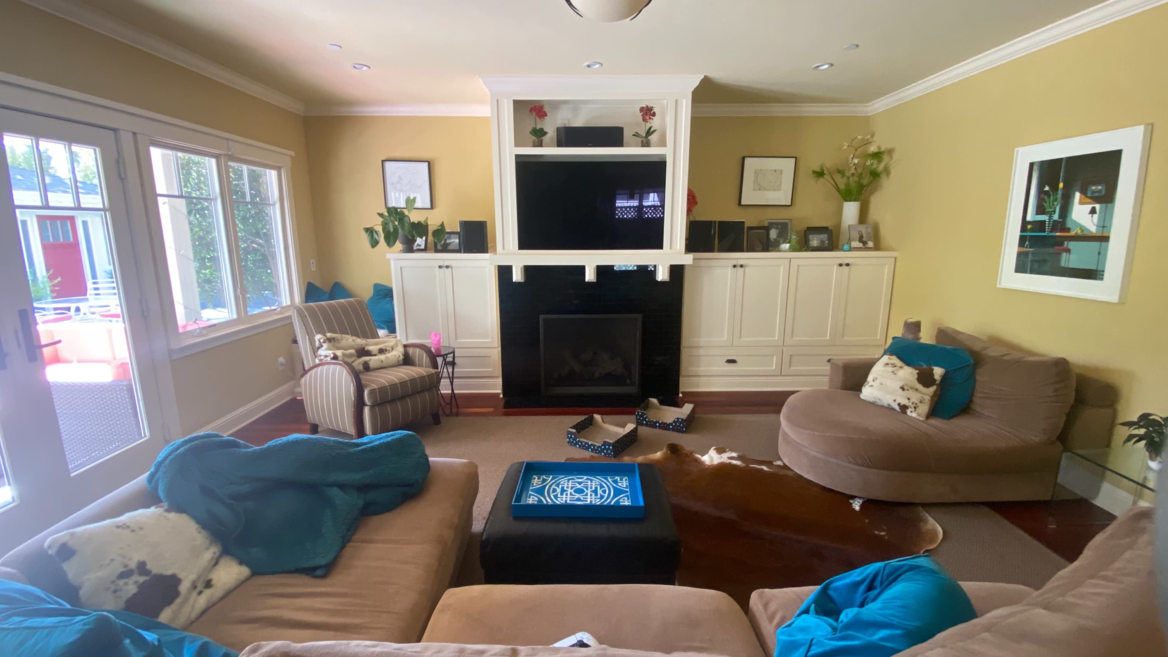 The image shows a living room with a beige sectional sofa, an armchair, a dark fireplace, and a wall-mounted television, surrounded by decorative plants and artwork.