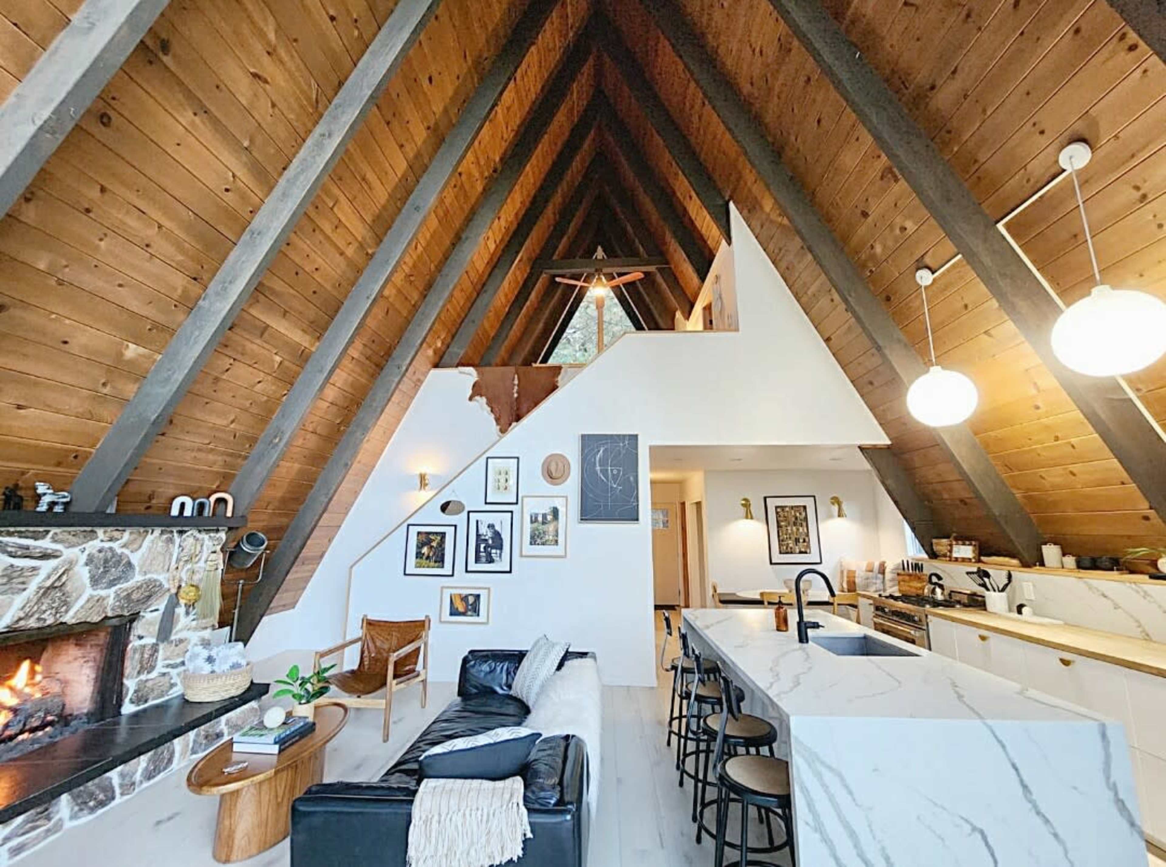 The interior of a spacious A-frame house features a stone fireplace, a modern kitchen with marble countertops, and large windows allowing natural light to fill the room.