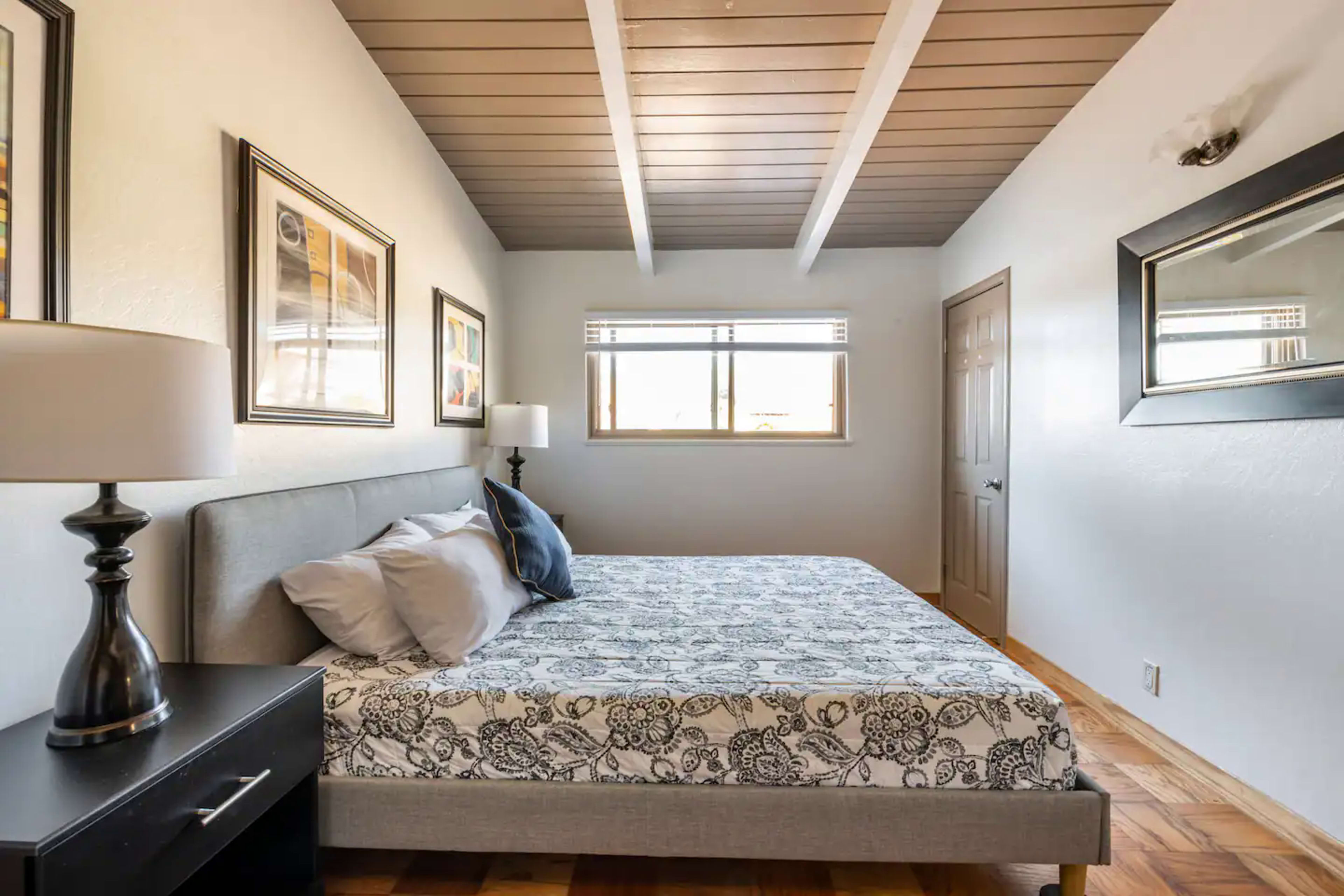 A bedroom with a bed, nightstand, and artwork on the walls, illuminated by natural light from a window.