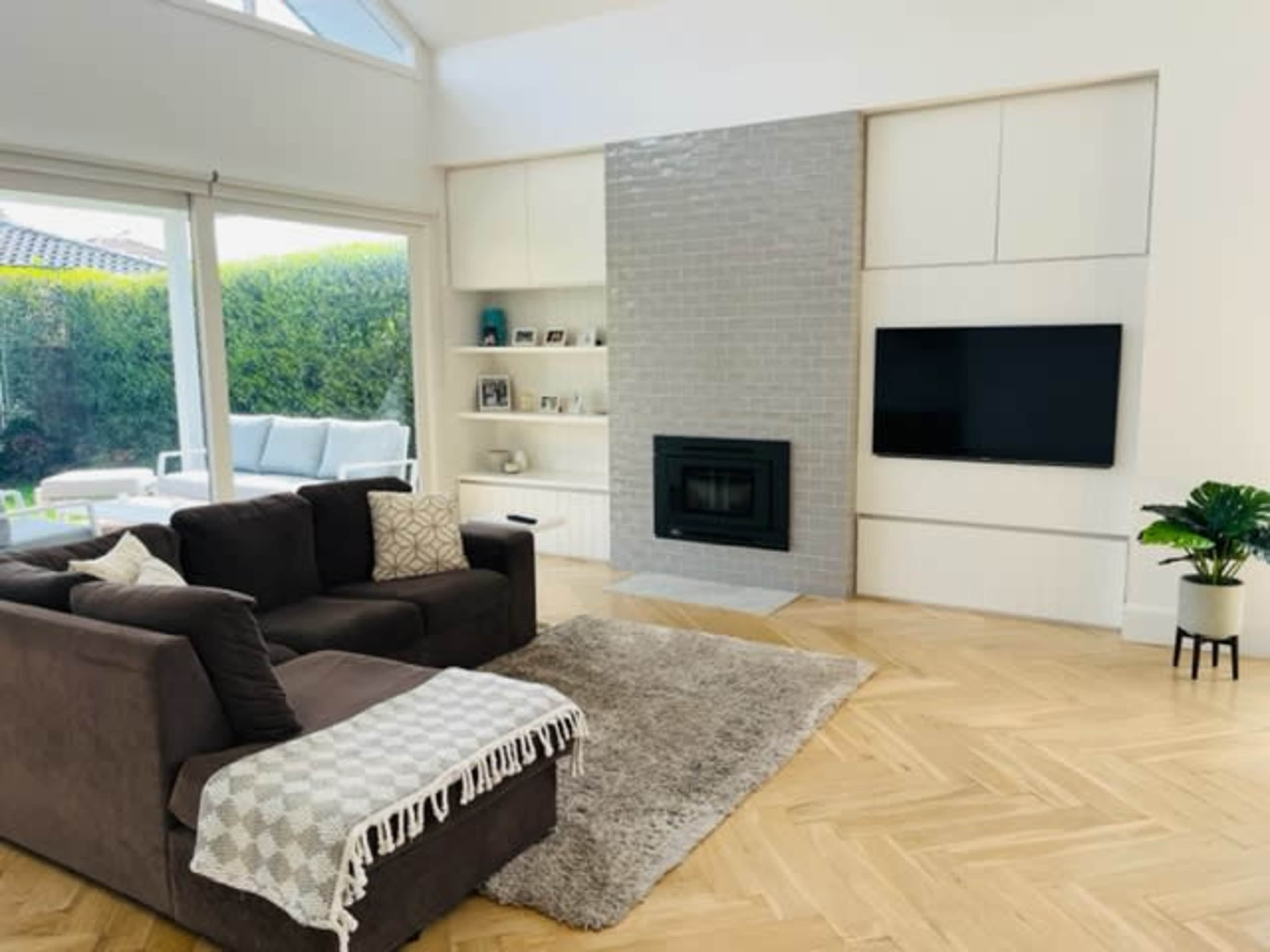 The living room features a gray sofa, a plush rug, a wall-mounted TV, and a fireplace, with large windows revealing an outdoor area.