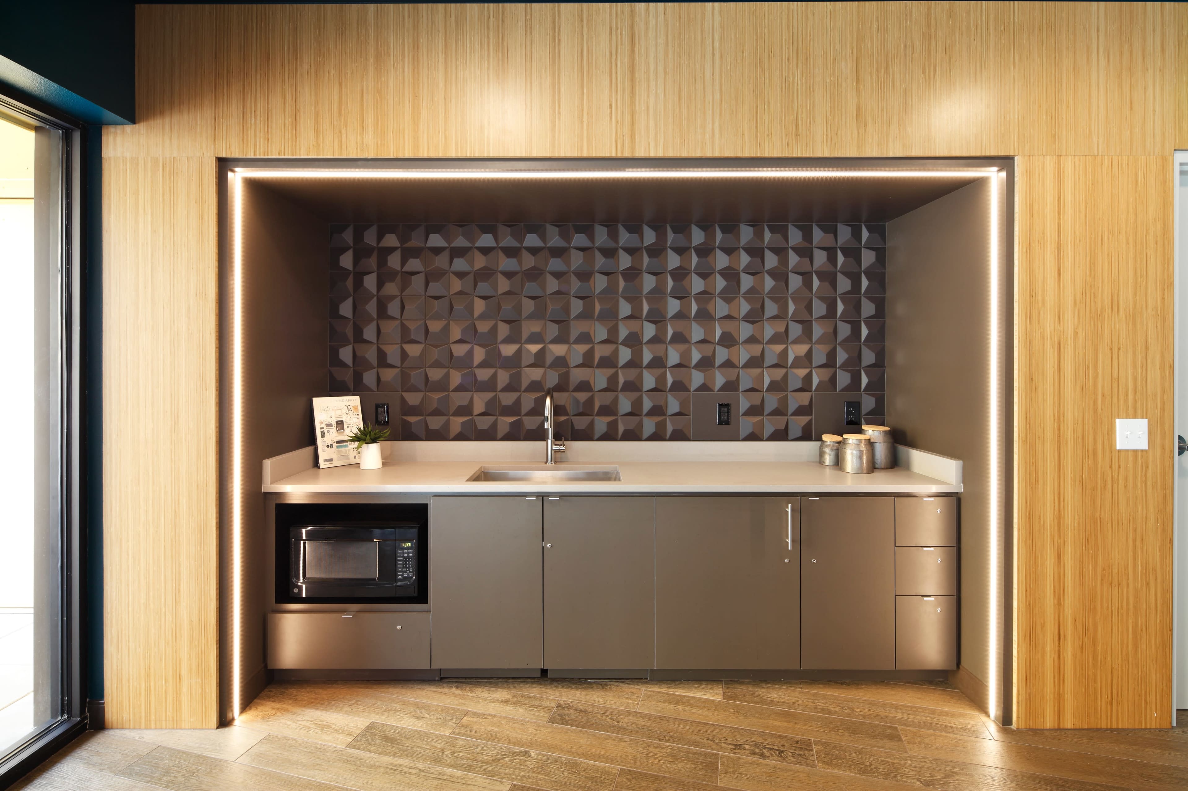 A modern kitchenette features a gray cabinetry with a sink, microwave, and geometric backsplash under soft lighting.