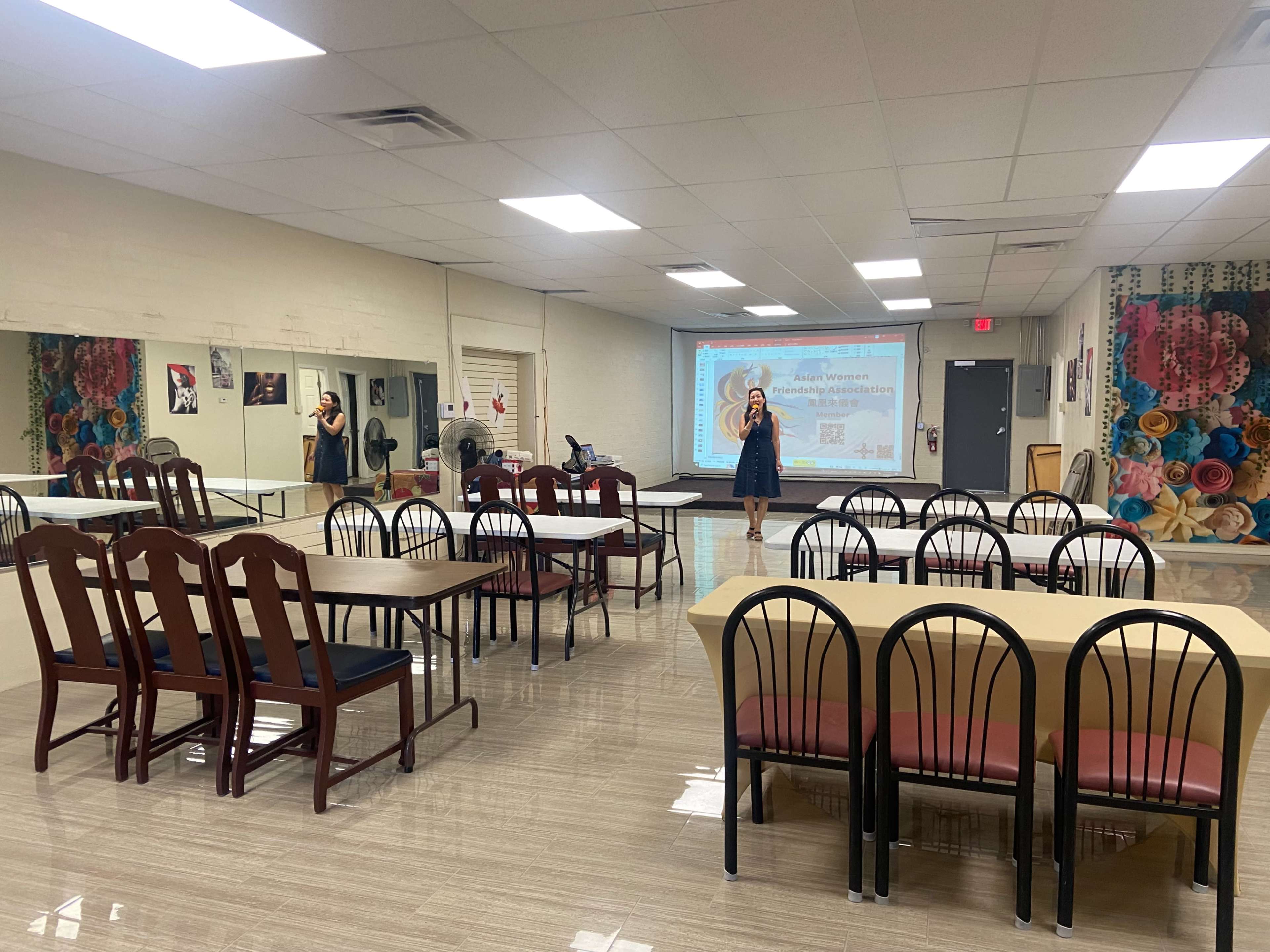 A spacious community hall features rows of tables and chairs, with a podium at the front and a large screen displaying a presentation.