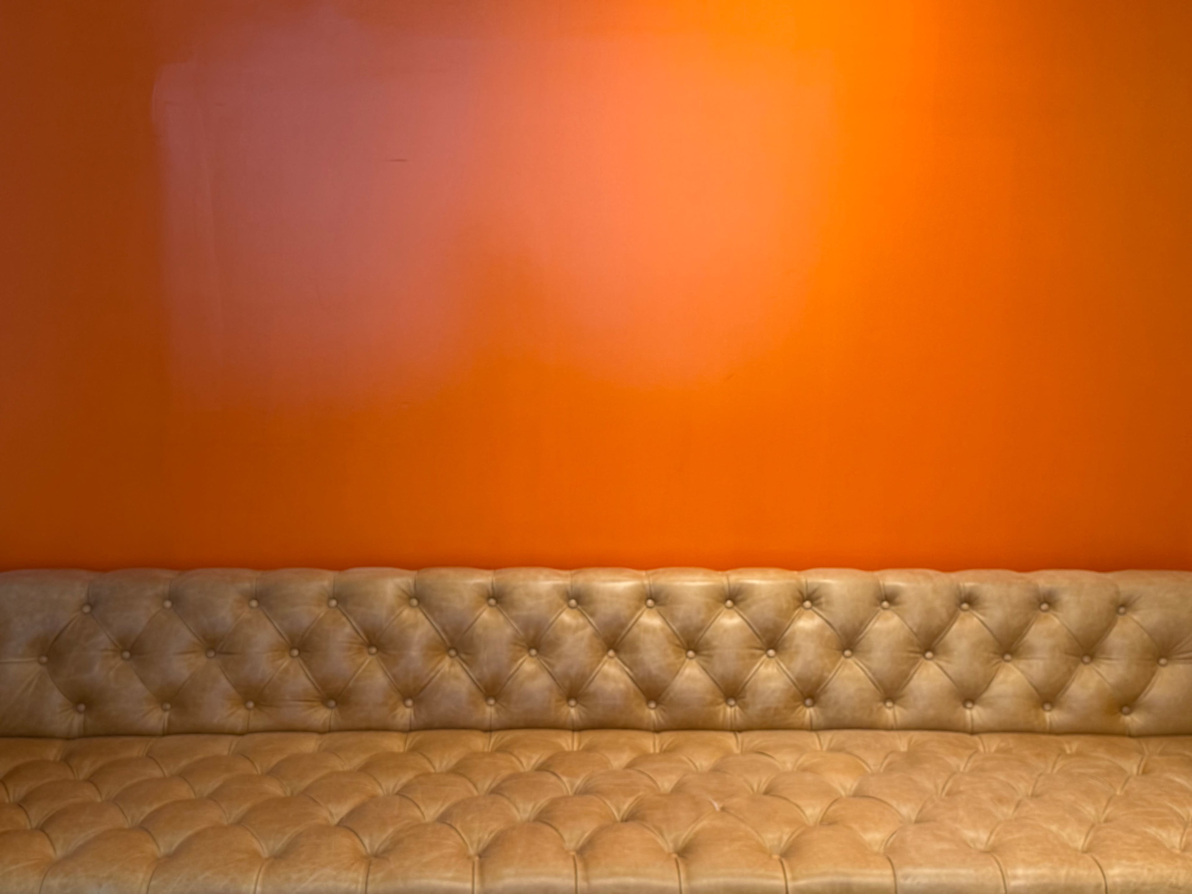 A tufted tan couch is positioned against a bright orange wall.
