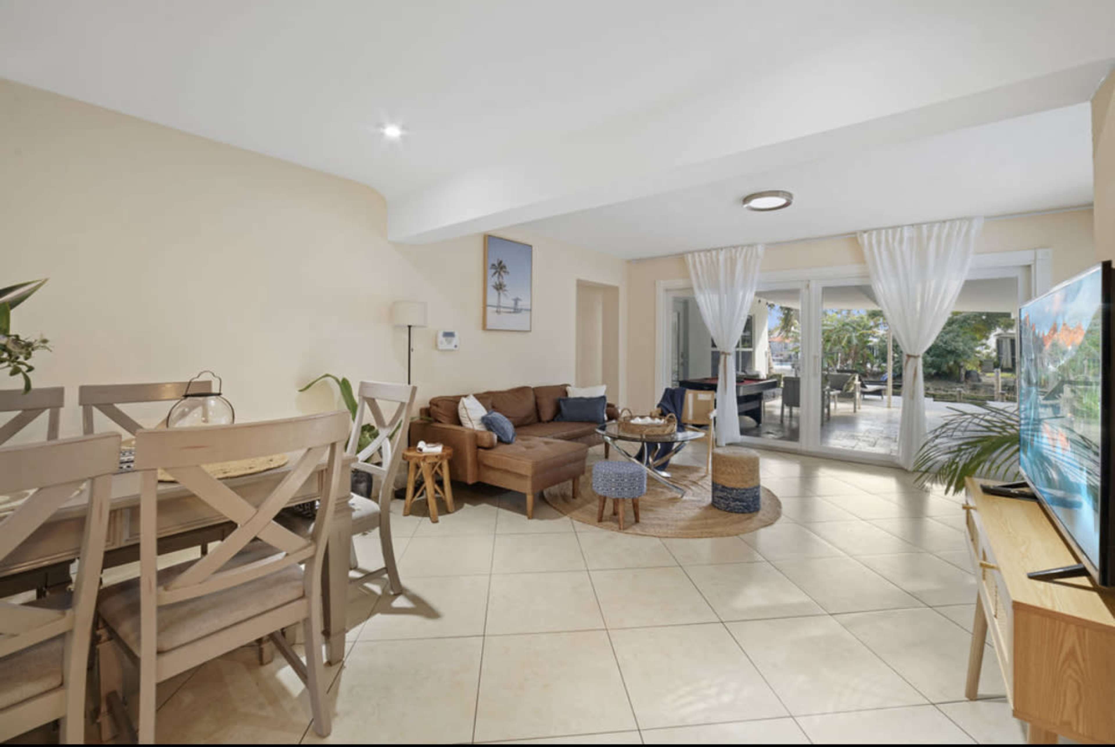 A light-colored, open living space featuring a brown sofa, a dining area with wooden chairs, and large glass doors leading to a patio.