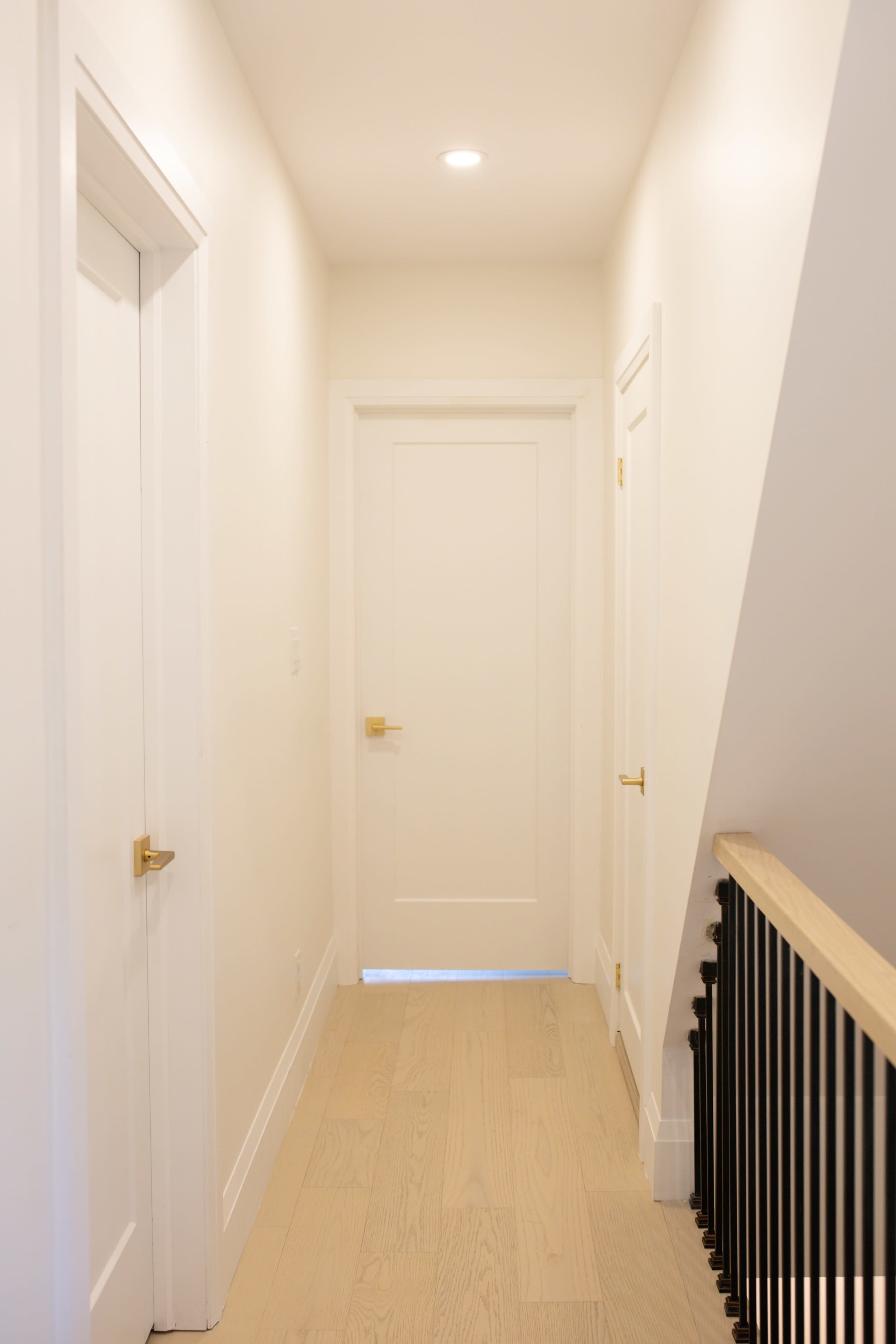 A narrow hallway features white walls, a light wooden floor, and doors on either side leading to rooms, with a railing visible on the right.